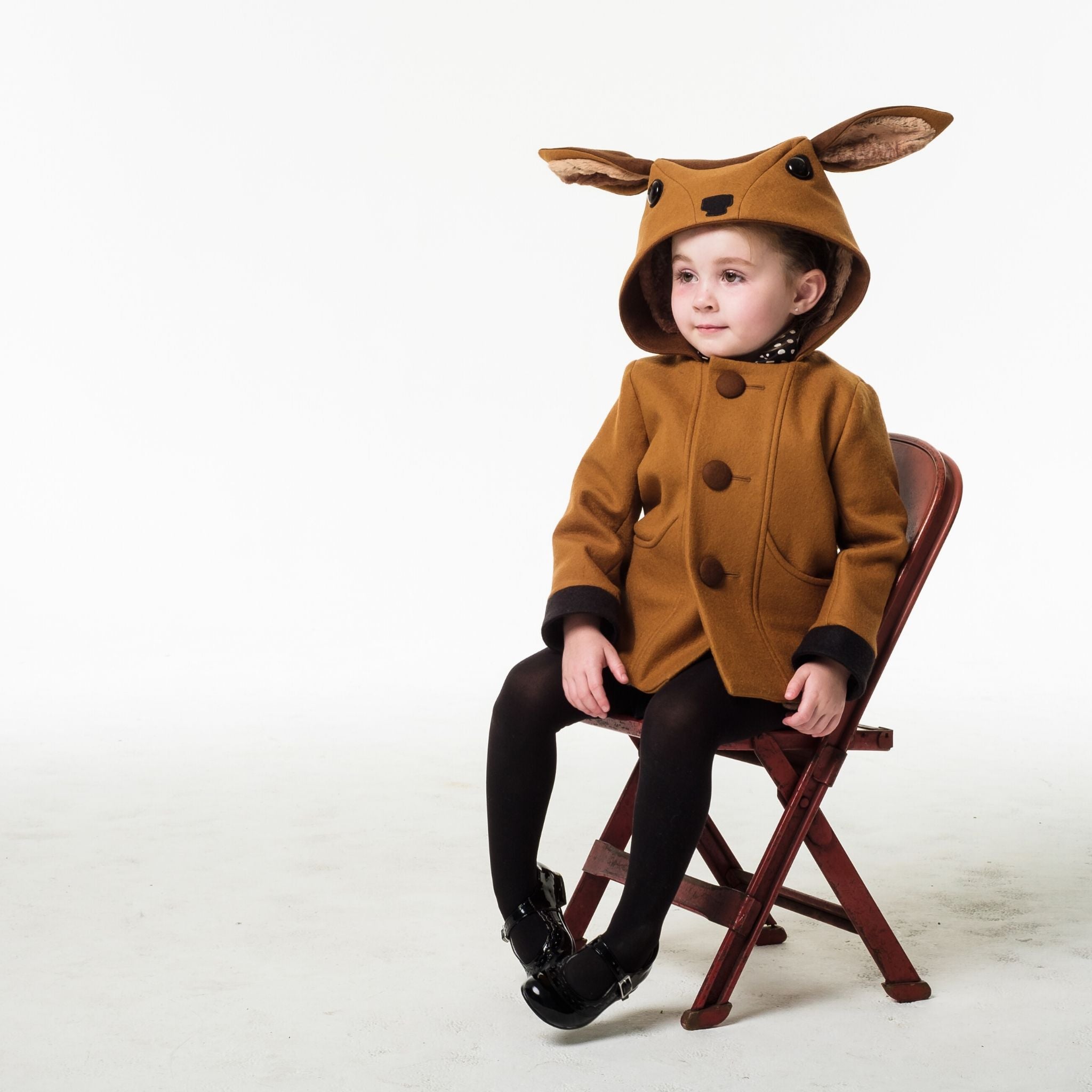 Child sitting in a Little Goodall Frolicsome Fawn Coat, featuring a playful hood with large ears and front pockets