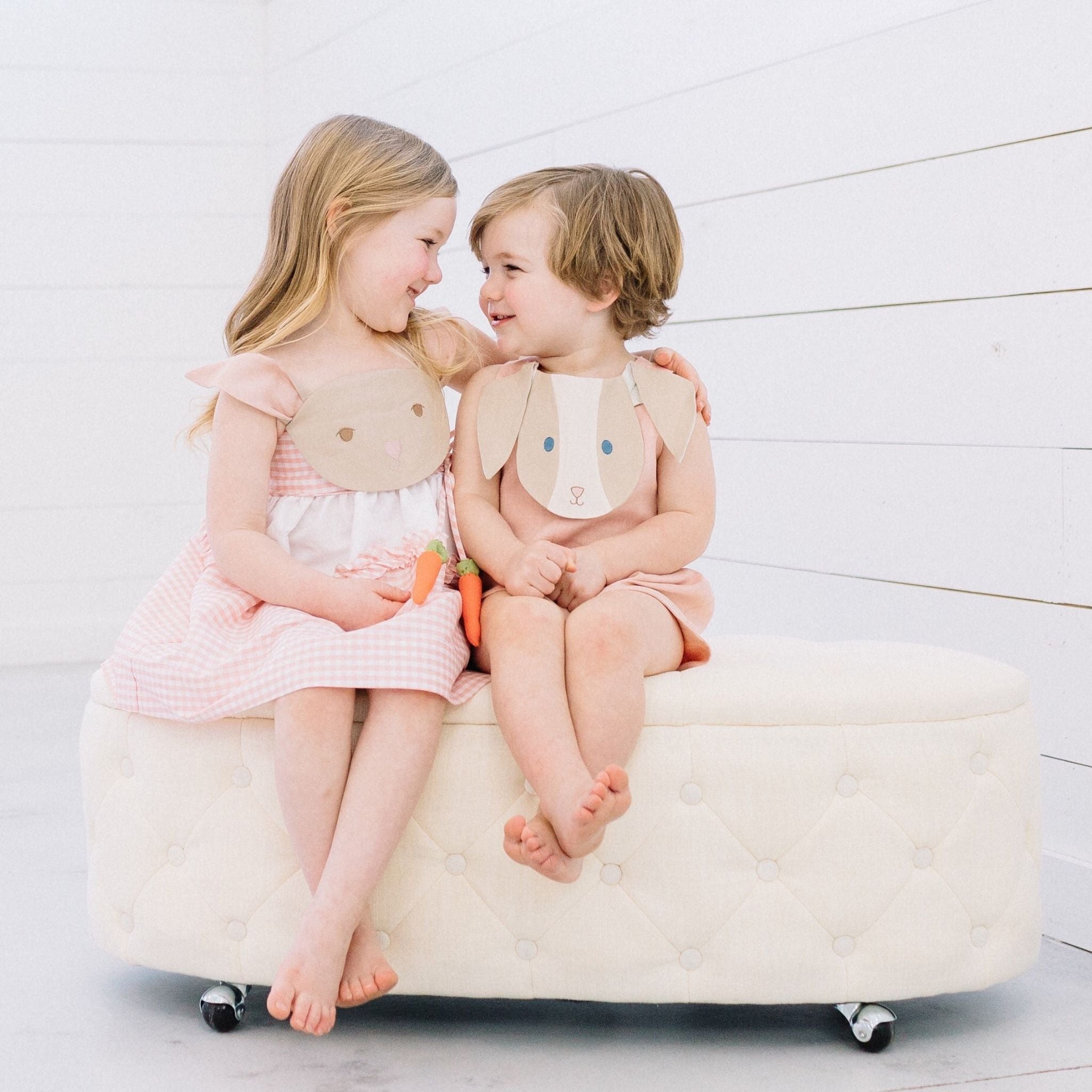 Two children smiling in coordinating Little Goodall bunny outfits—a pink gingham Bunny Love Dress and Bunny Face Romper with floppy ears and carrot details