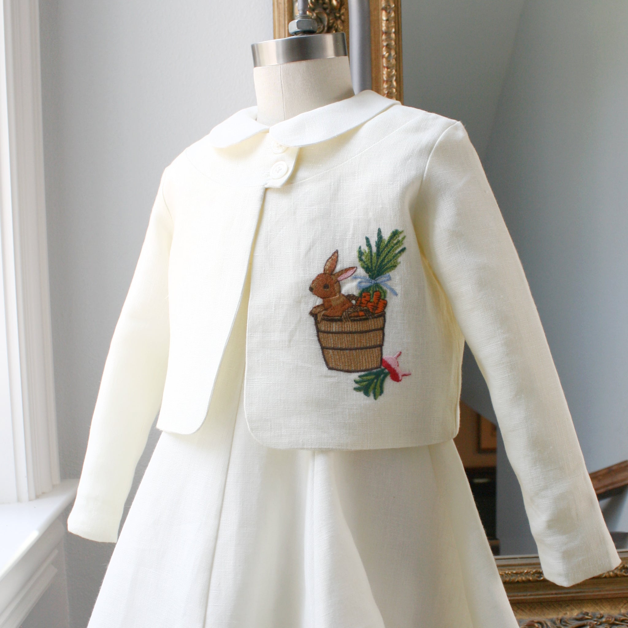 Ivory linen bunny jacket by Little Goodall on dress form, featuring embroidered bunny in a bushel of carrots and radishes
