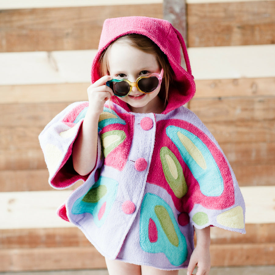 Child wearing a colorful butterfly hooded cape with pink, blue, and green wing details, smiling and adjusting heart-shaped sunglasses.