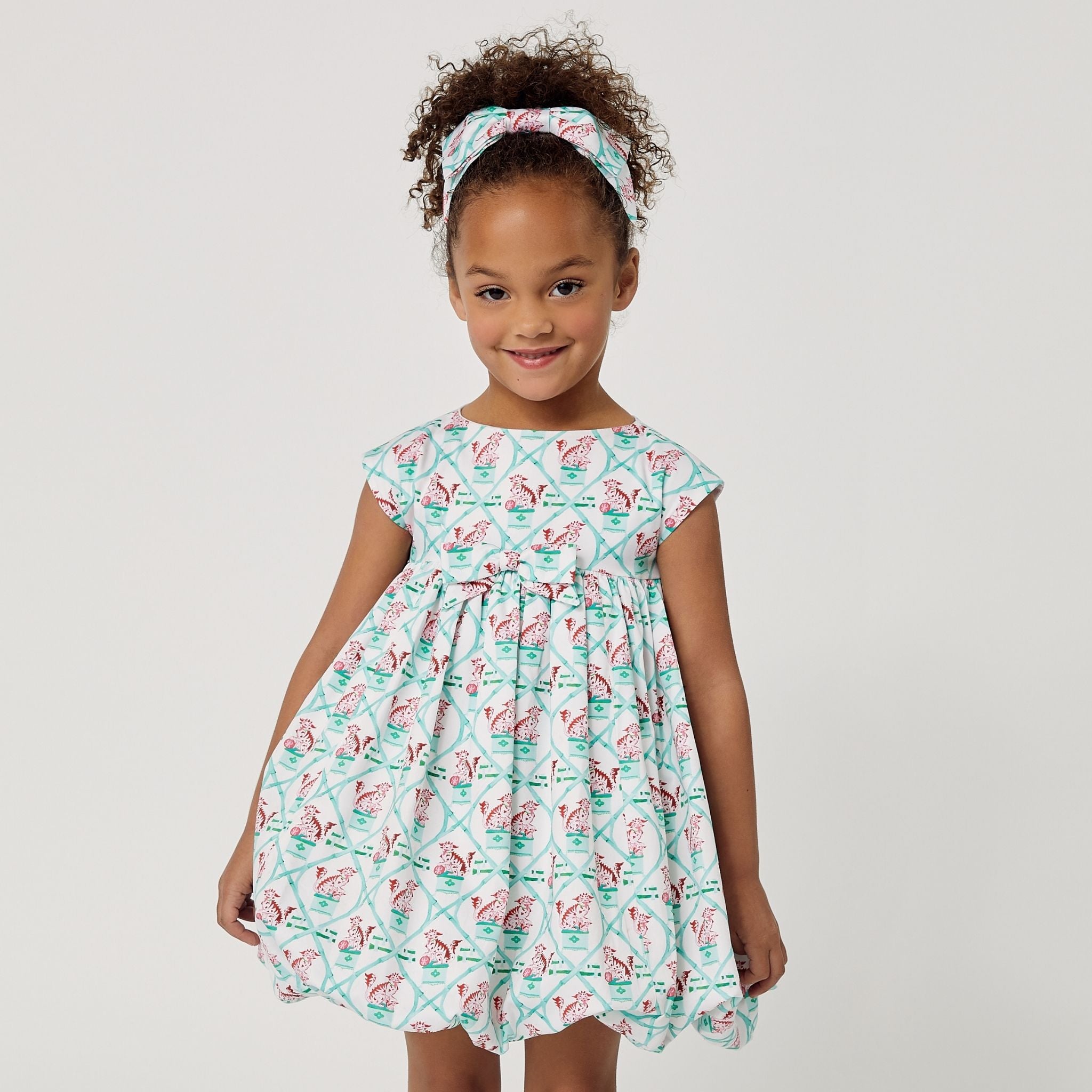 Girl wearing the Carrot and Dot Dress in mint with a bamboo trellis print, cap sleeves, and a gathered balloon skirt,with her hands at sides