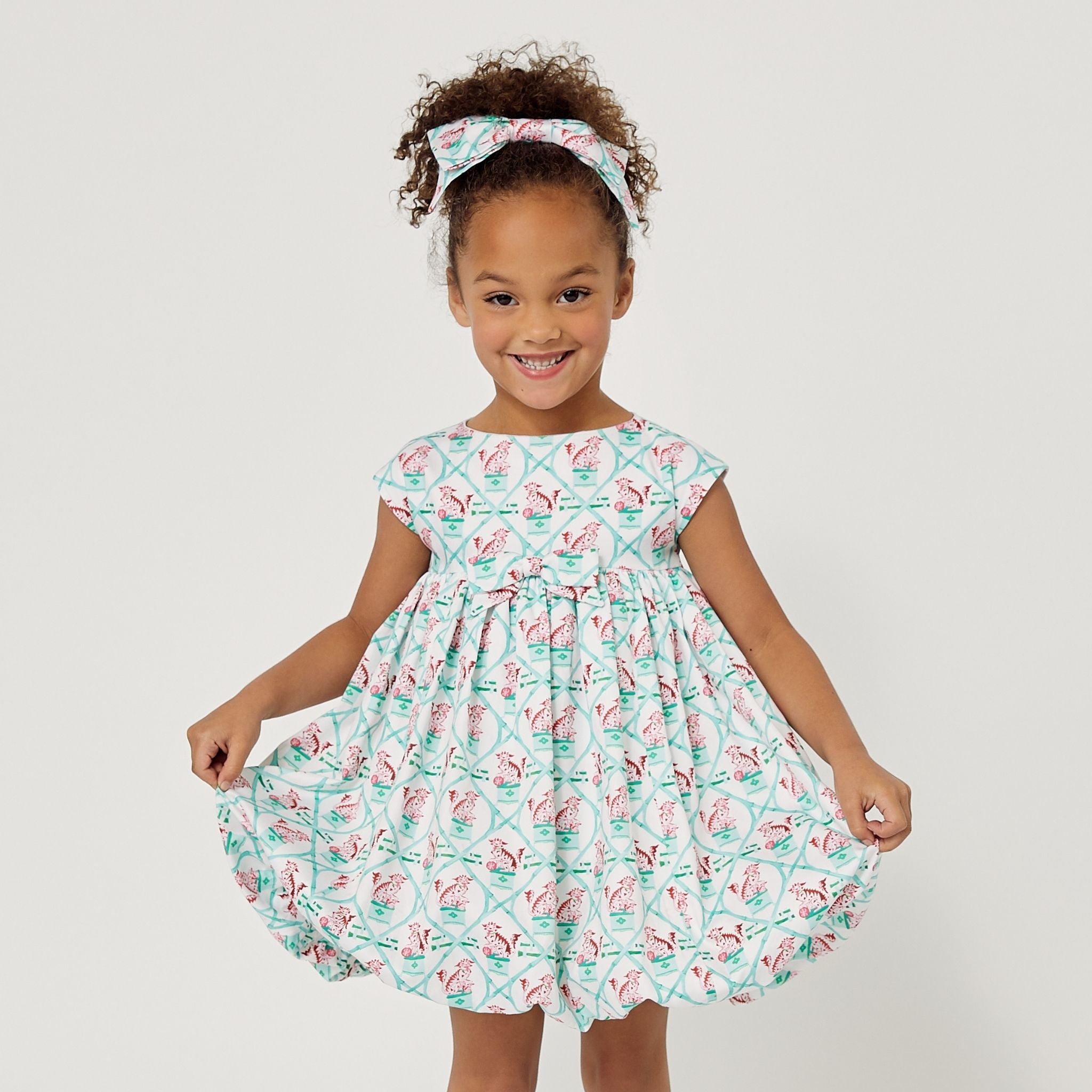 Girl in the Carrot and Dot Dress in mint & plum with a bamboo trellis print, cap sleeves, and a balloon skirt, holding the sides of her dress