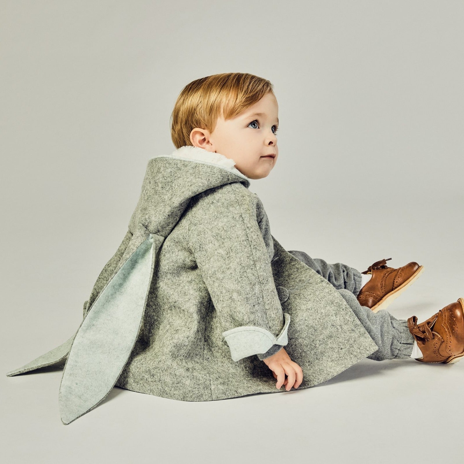 Seated child in a Little Goodall grey bunny coat, showcasing floppy ears with a light blue lining.