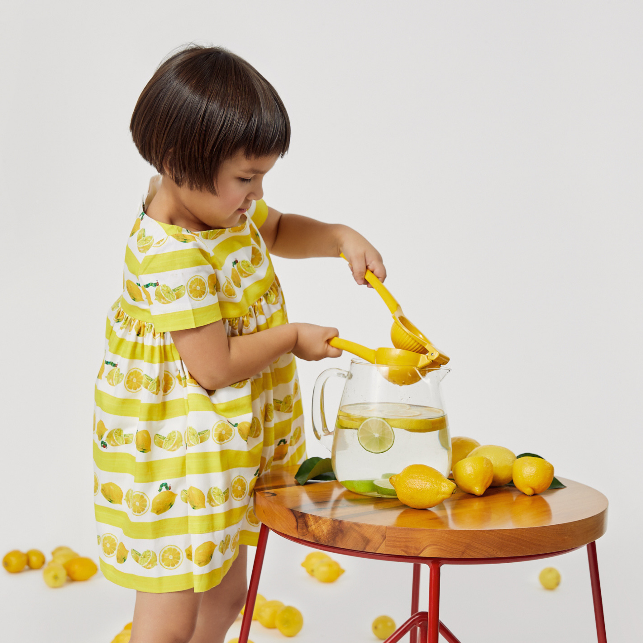 A child in a lemon-print dress squeezes a lemon into a glass pitcher on a wooden table with fresh lemons and limes.