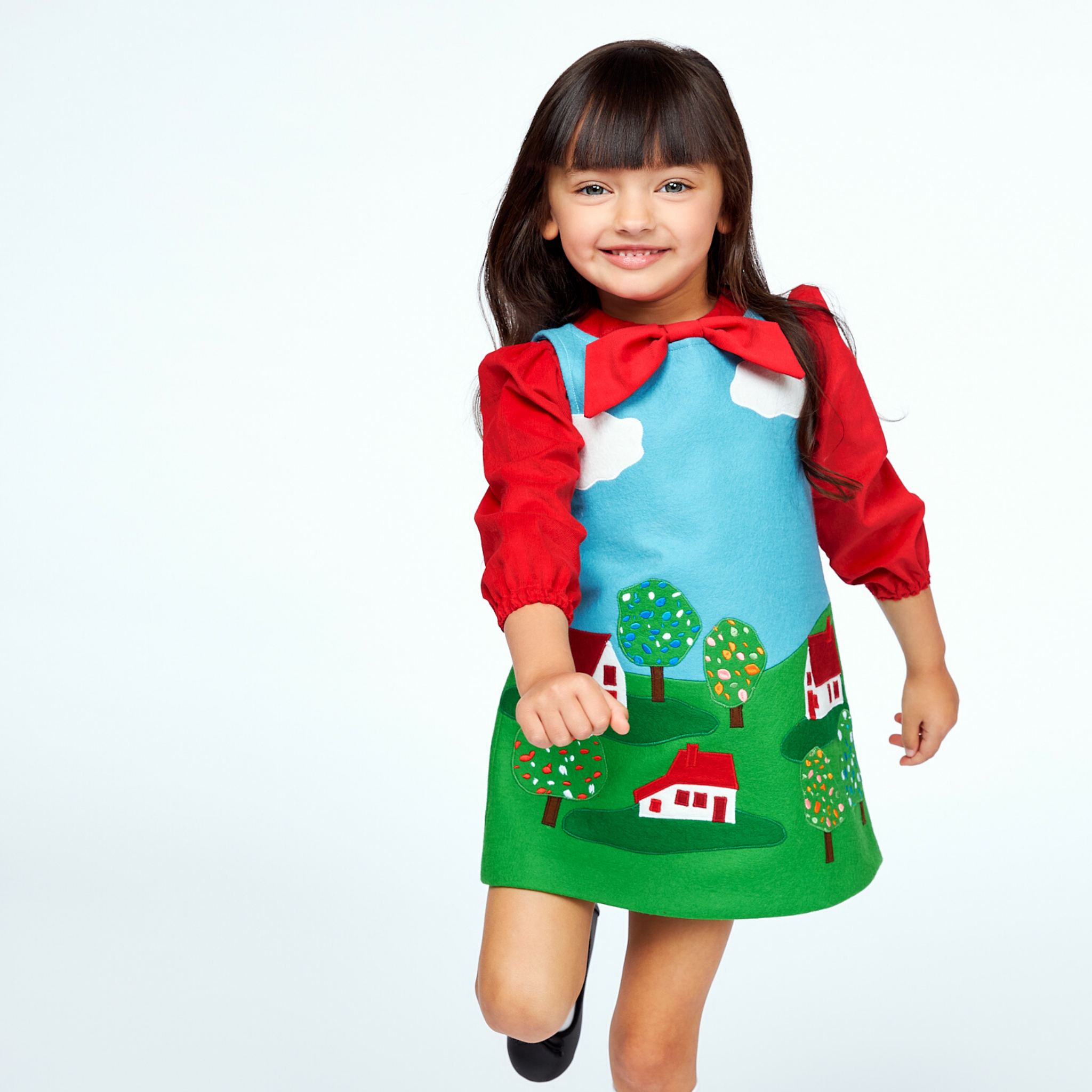 Adorable toddler in a red, blue, and green jumper dress with cloud and landscape motifs, perfect for imaginative play.