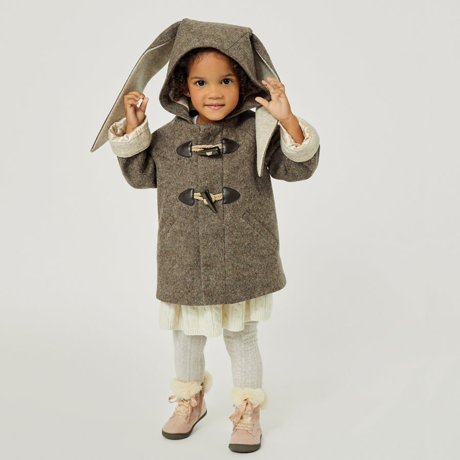 Smiling toddler wearing a wool Flopsy Bunny Coat with floppy ears, toggled front, and a cozy hood, playfully lifting the bunny ears