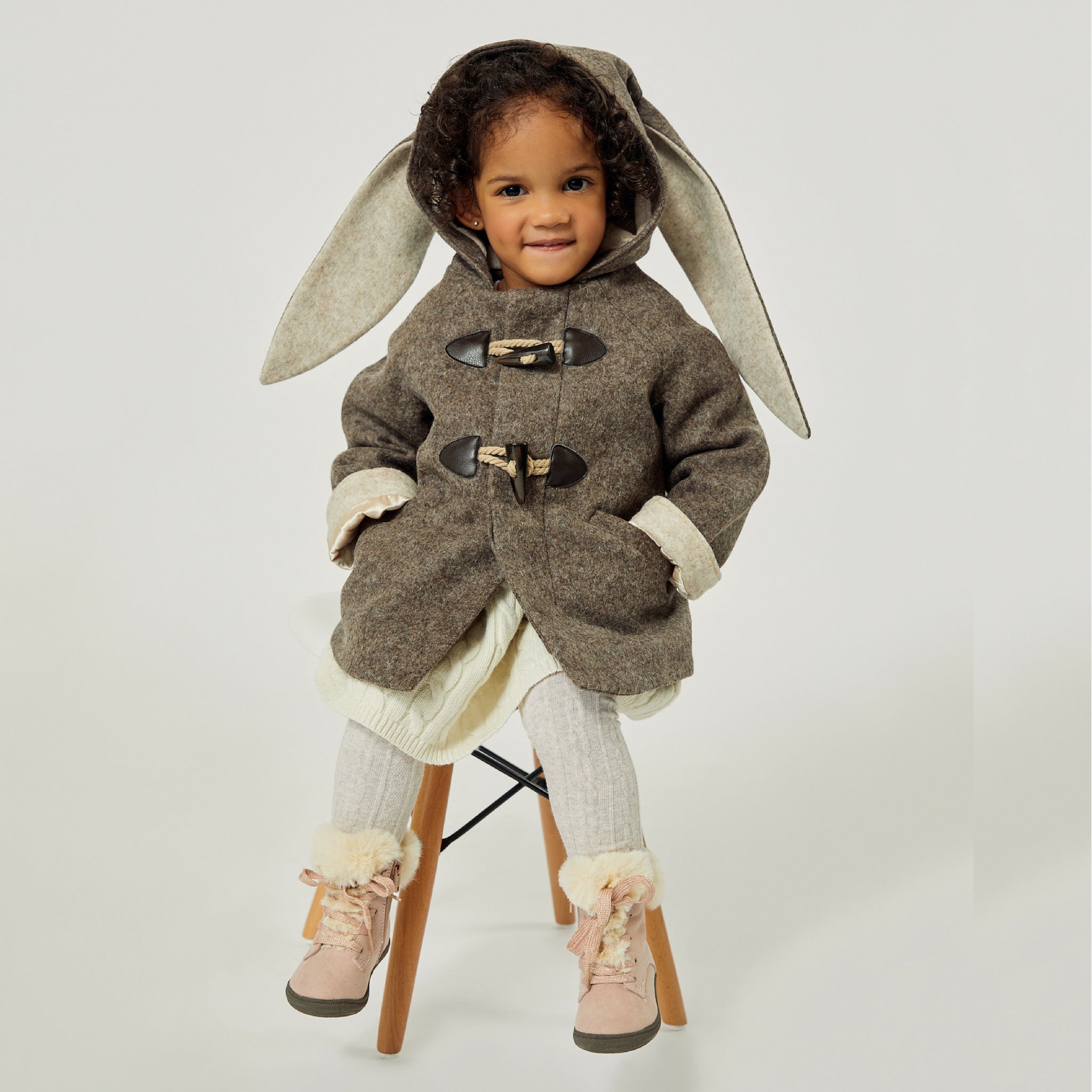 girl sitting on a stool wearing the Little Goodall Flopsy Bunny Coat, a cozy wool jacket with floppy bunny ears and toggle closures, perfect for winter