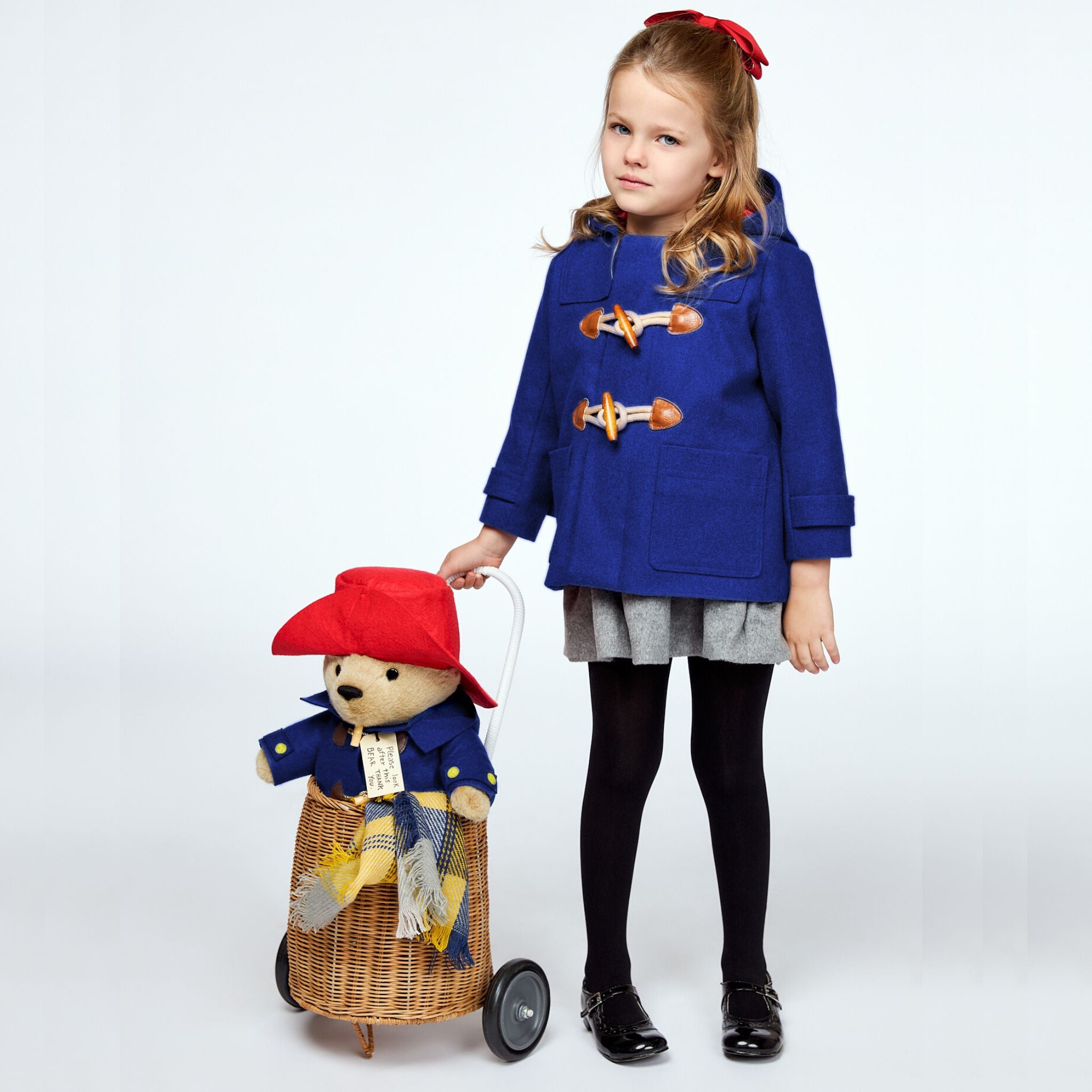 girl in a blue wool duffle coat with wooden toggles, standing beside a wicker cart carrying a Paddington Bear plush toy in a matching coat and red hat