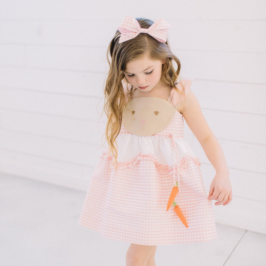 Girl wearing Little Goodall Pink Gingham Bunny Love Dress with matching hair bow, bunny face bodice, and dangling carrot details.