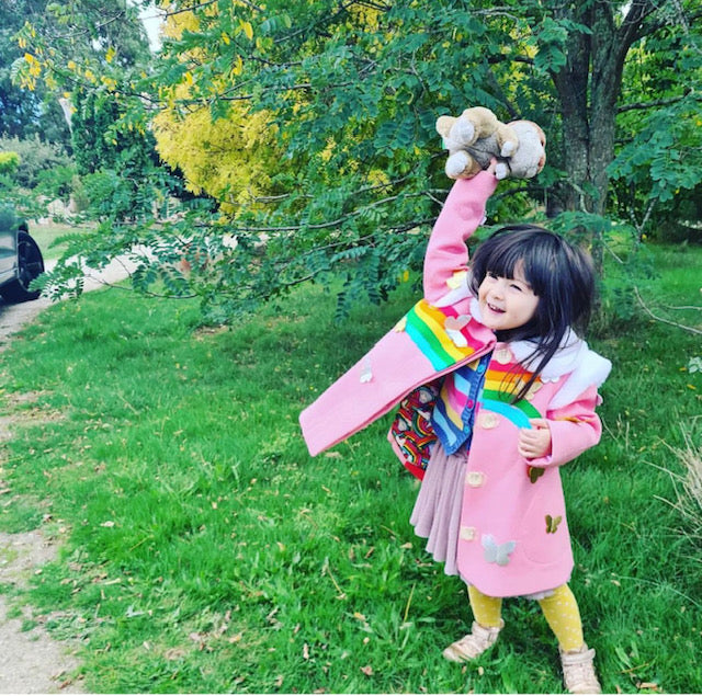 Smiling girl in a pink wool coat with rainbow appliqués and butterflies, playing outside with a stuffed bunny, coat lining peeking through