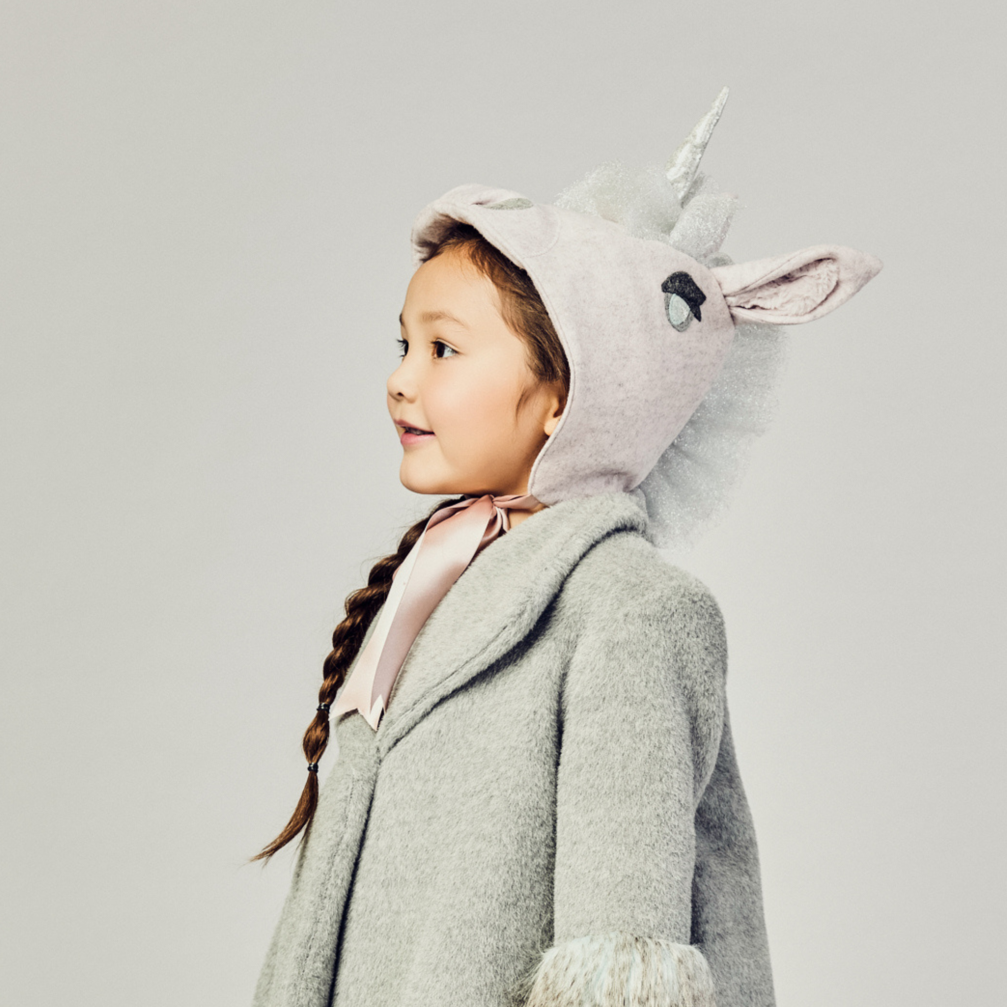 Young girl wearing a pale pink wool unicorn hat with a silver mane and satin ribbon ties, styled with a cozy grey winter coat