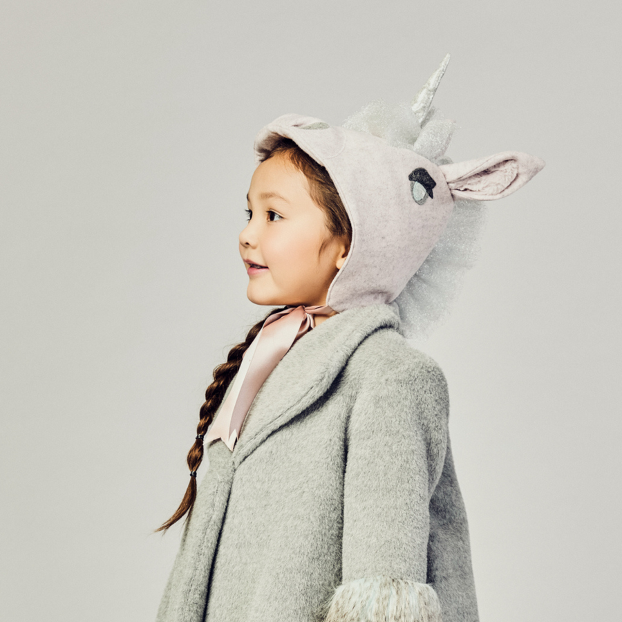 Young girl wearing a pale pink wool unicorn hat with a silver mane and satin ribbon ties, styled with a cozy grey winter coat