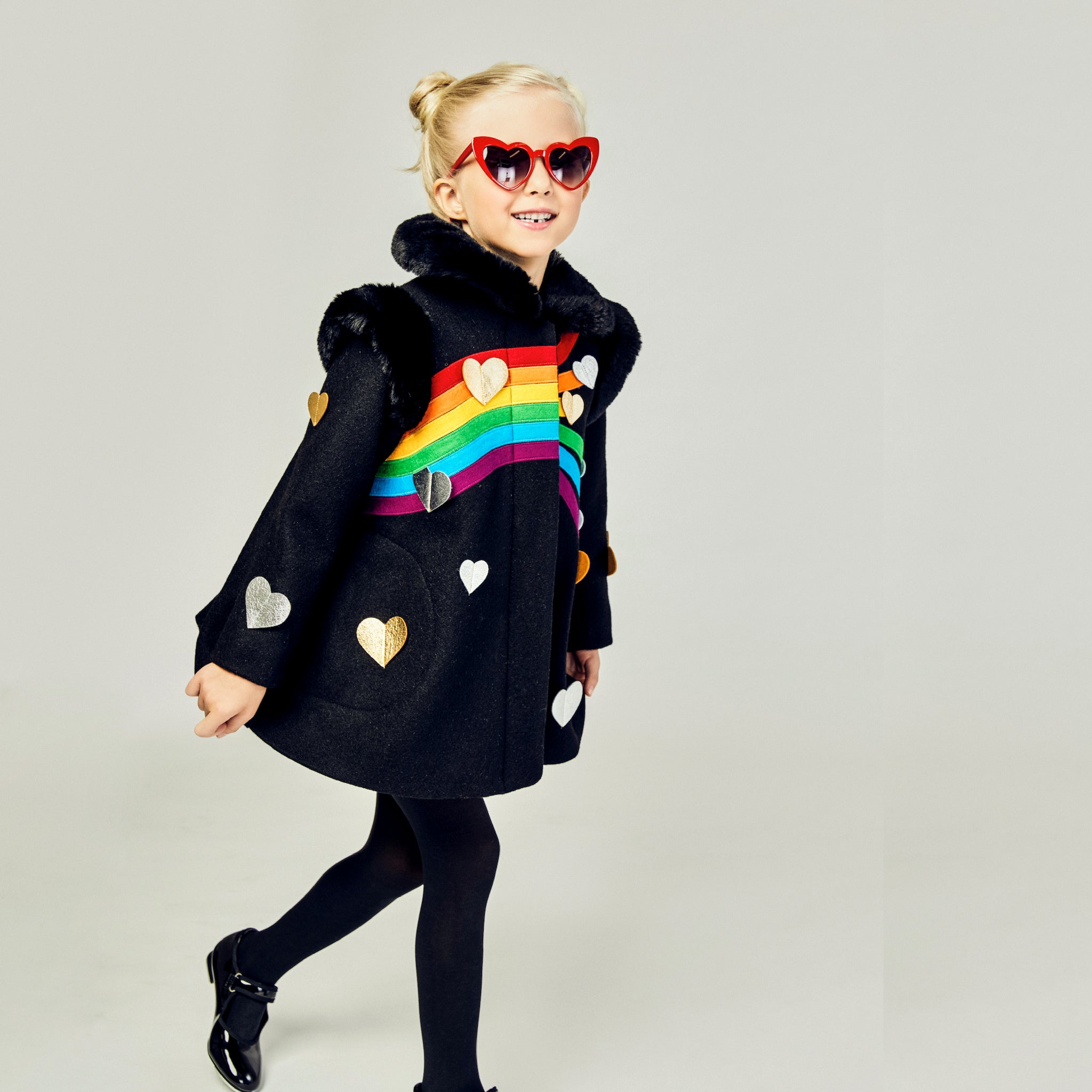 Girl modeling black coat with wearing black tights and shoes and her hair in two buns with red heart sunglasses on.
