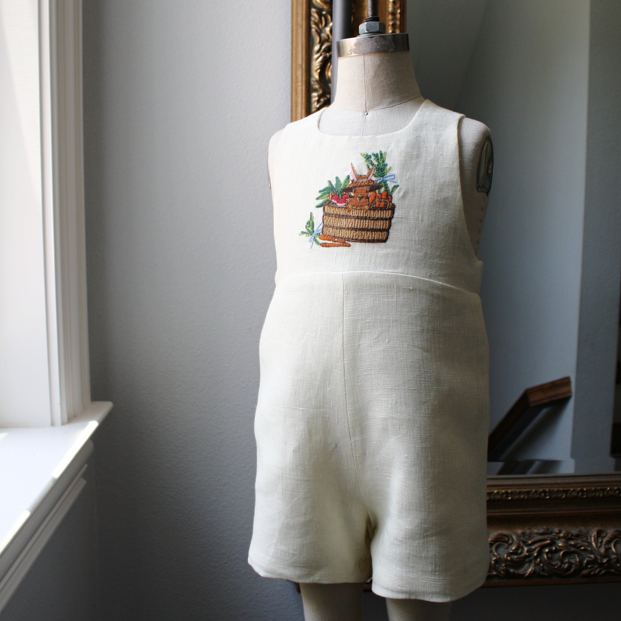 Ivory linen bunny romper with embroidered basket, displayed on a dress form. Classic gender-neutral baby outfit by Little Goodall