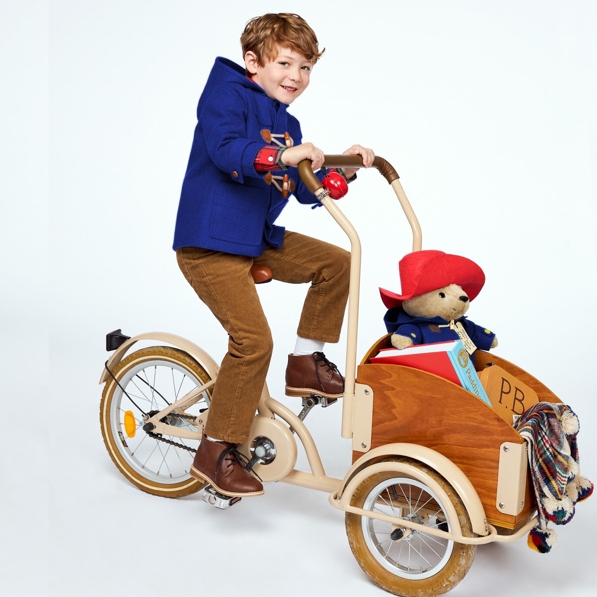 Child wearing a royal blue-inspired wool coat with wooden toggles and a hood, riding a tricycle with a Paddington Bear plush toy in a wooden cargo box