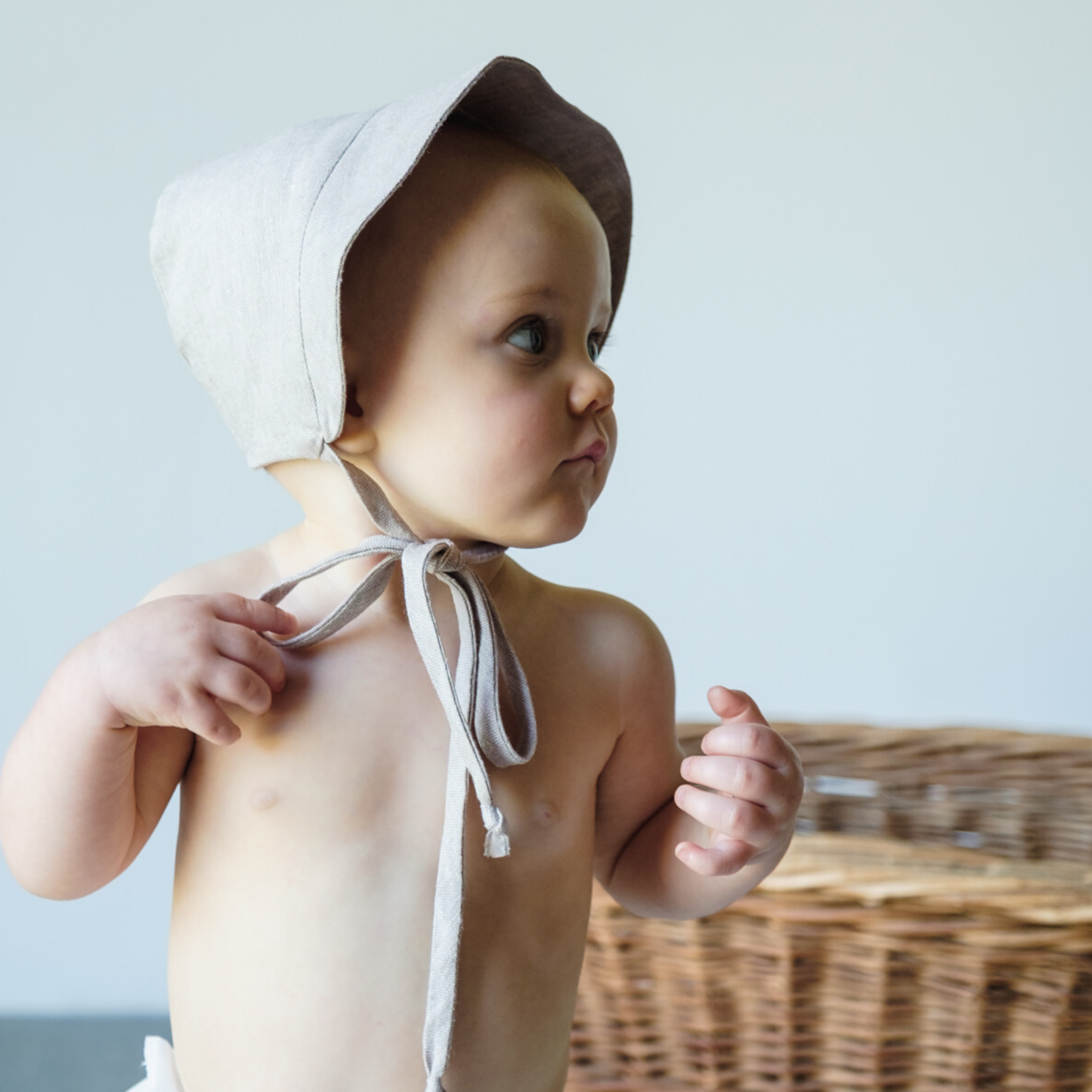 Baby in a wheat-colored linen sun bonnet with a wide brim and chin ties, shown from the side