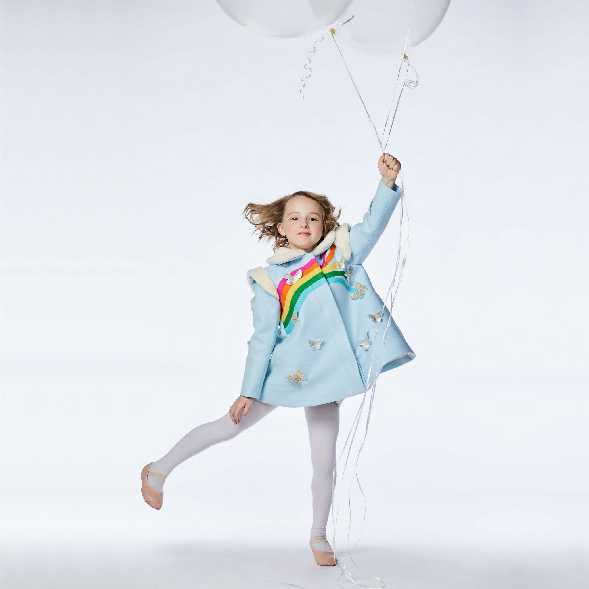 Girl in a sky blue wool coat with rainbow and butterfly details, holding white balloons while posing playfully