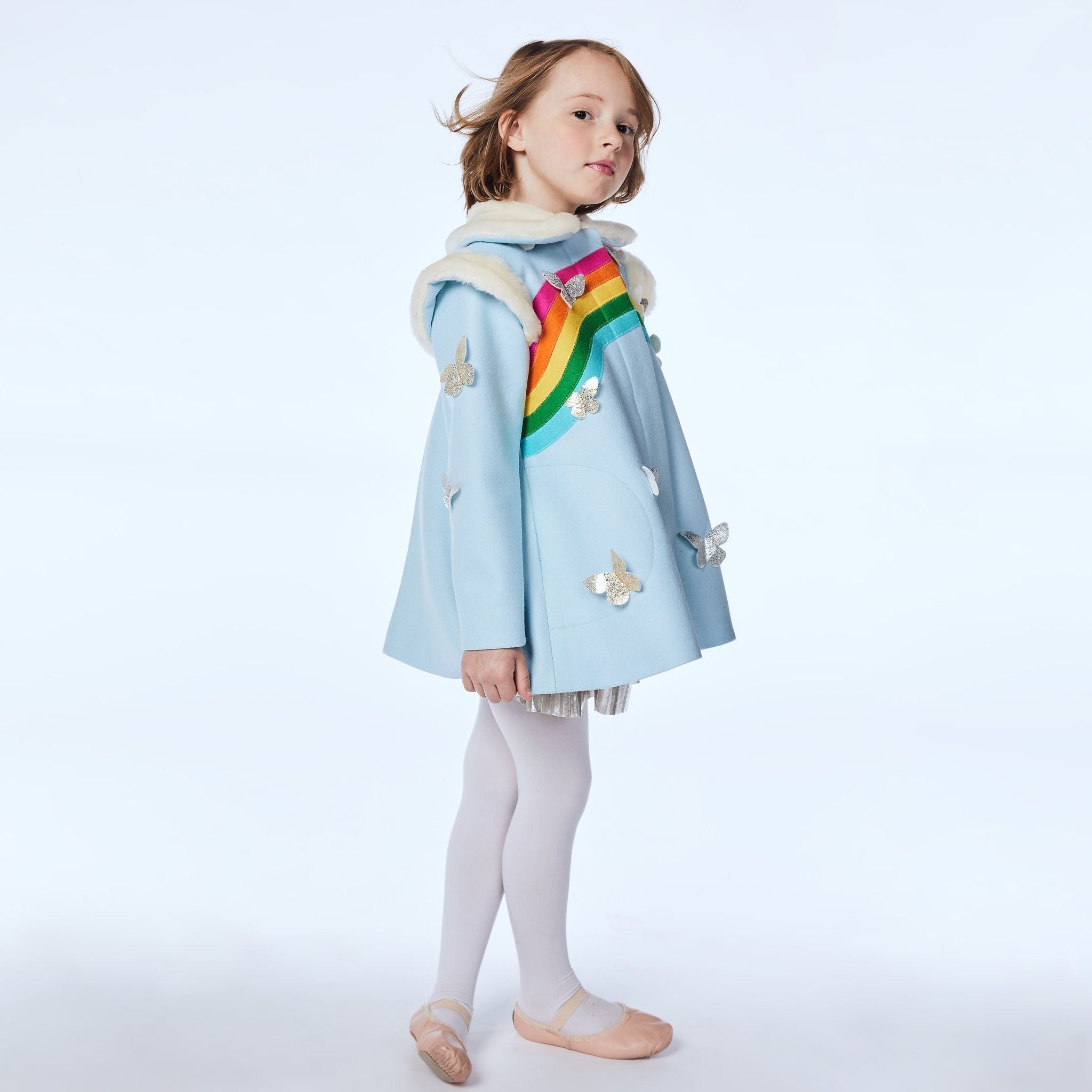 A  girl poses in the Rainbow Butterfly wool coat, a sky-blue dress coat with a faux fur collar, rainbow detailing, and metallic butterfly appliqués