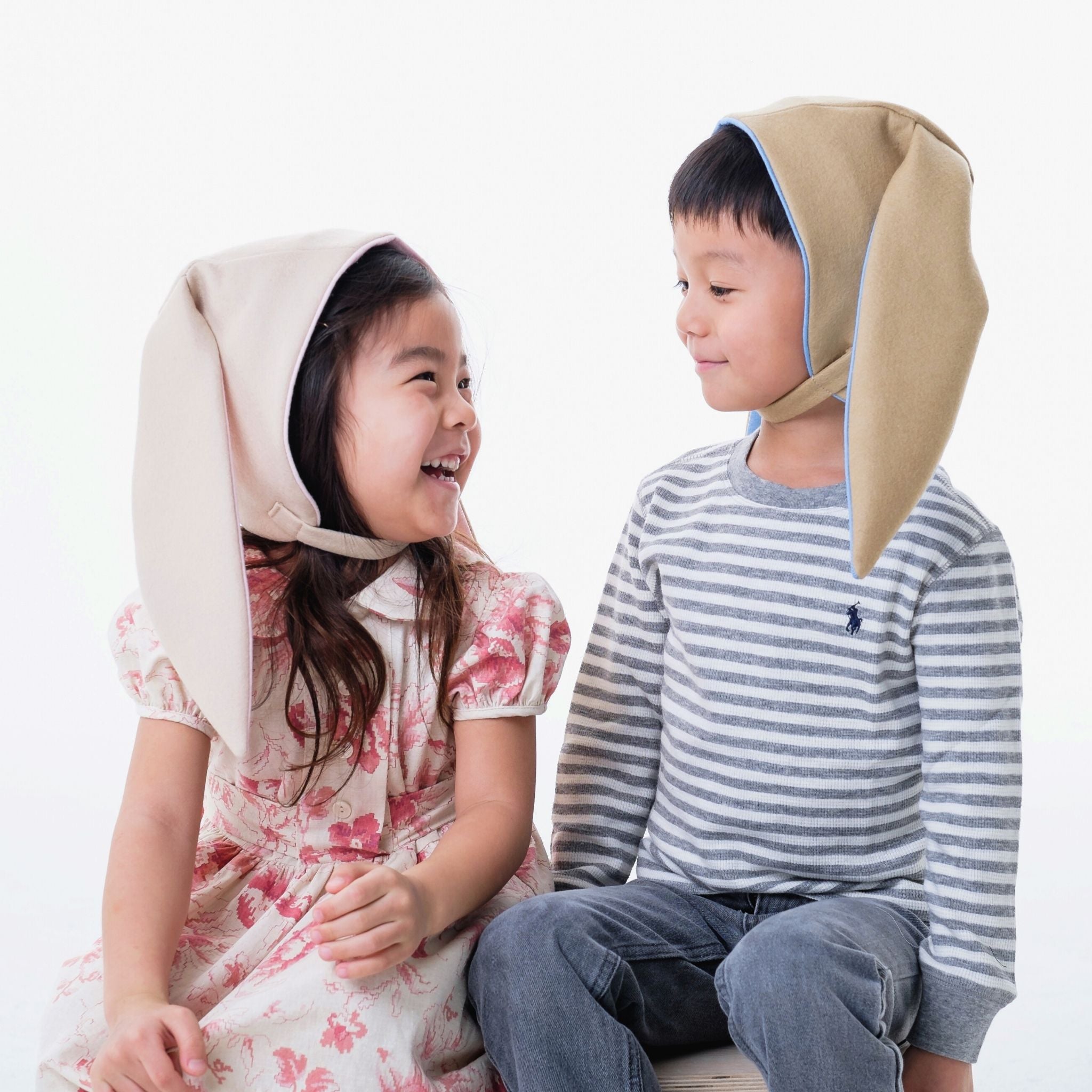 children wearing cozy beige hats with blue linings; the design features playful bunny-inspired details, made from 100% Merino wool and fully lined with soft fleece for warmth.