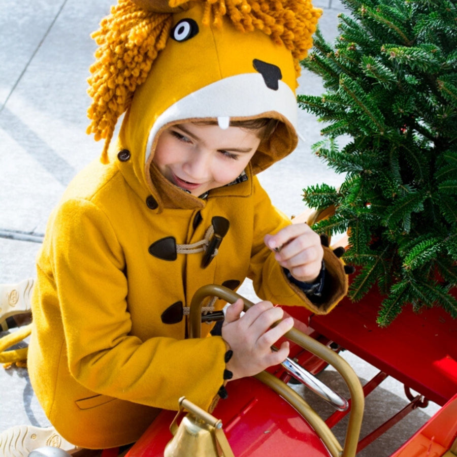 Child in Little Goodall Luxe Lion Coat with hood up, showing lion face design, riding a red sled with a Christmas tree. Warm and playful kids' jacket