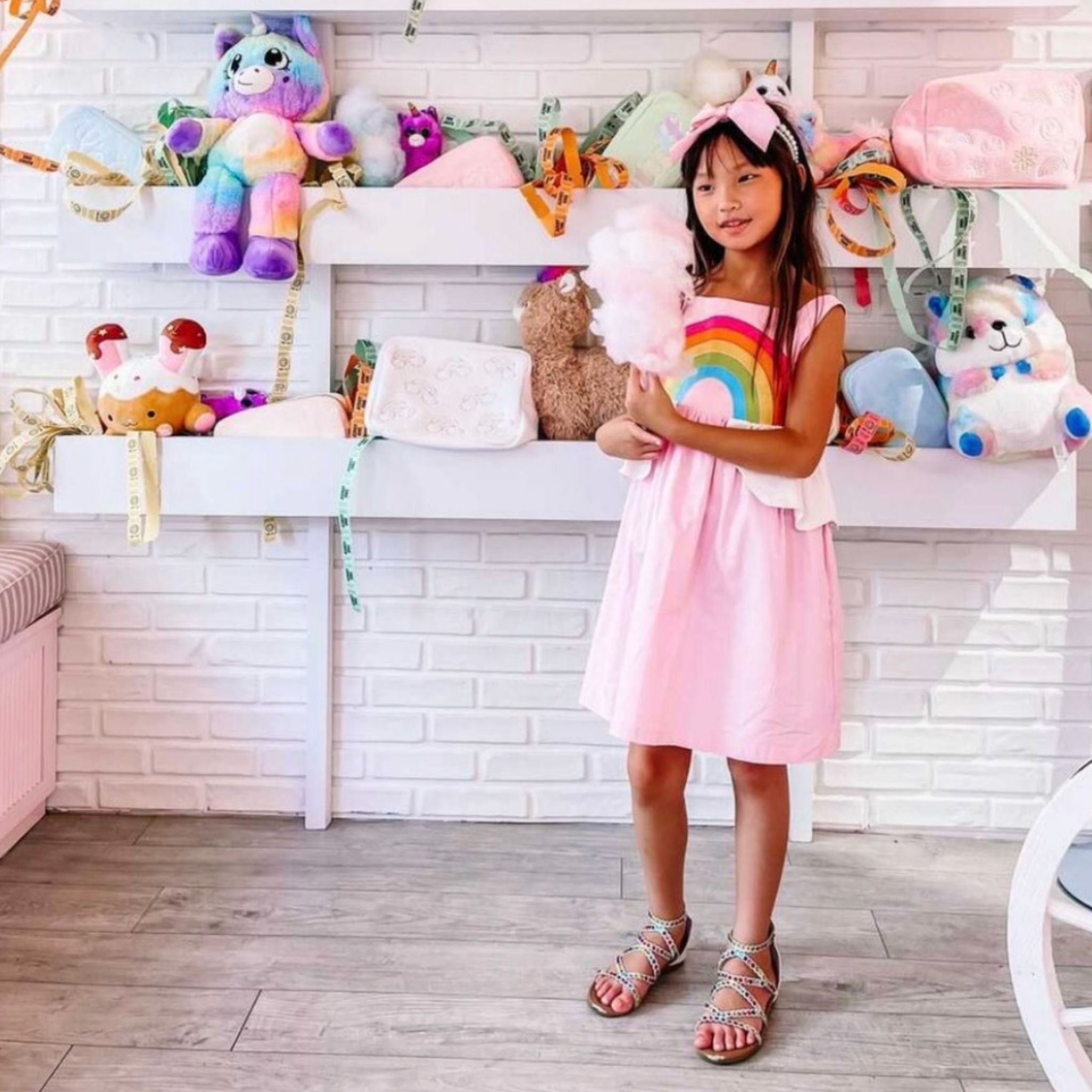 Girl in pink rainbow sundress holding cotton candy in a toy-filled boutique, wearing strappy sandals and a unicorn headband