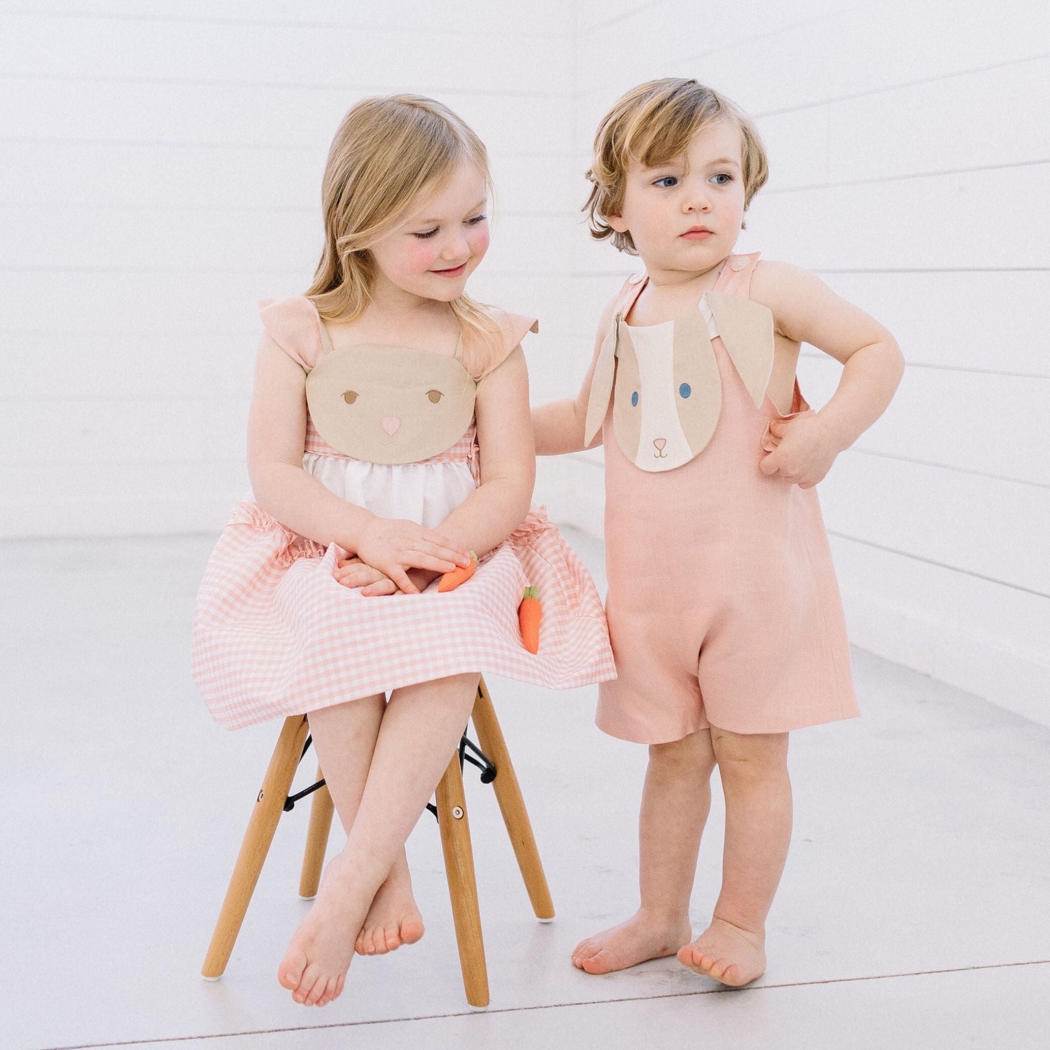Two children wearing Little Goodall bunny-themed outfits—pink gingham Bunny Love Dress with carrot details and coordinating pink Bunny Face Romper
