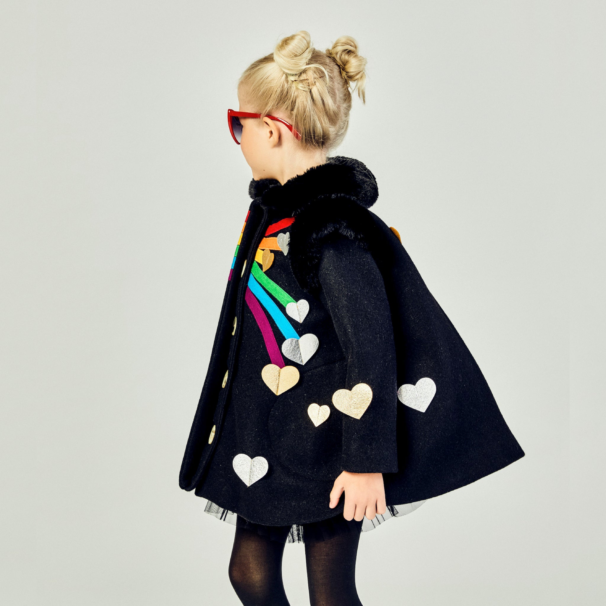 Girls' Queen of Hearts coat in black wool with rainbow stripes, metallic heart appliqués, and faux fur collar, shown from a side angle.