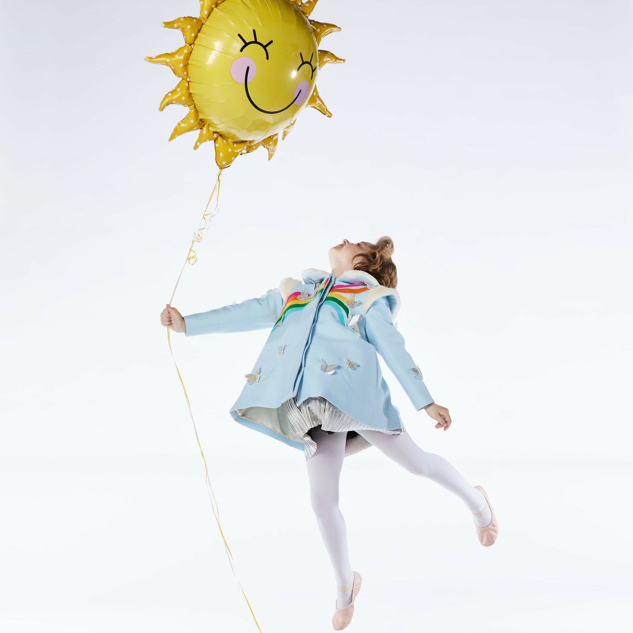 A girl in the Rainbow Butterfly wool coat, white tights, and ballet slippers leaps joyfully, holding a yellow sun balloon 