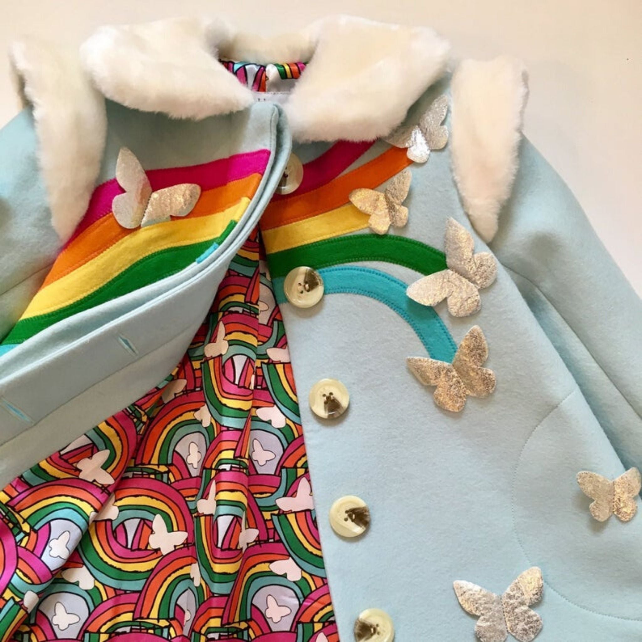 Close-up of Rainbow wool coat, showcasing the soft faux fur collar, vibrant rainbow appliqués, metallic butterflies, and colorful rainbow-print lining
