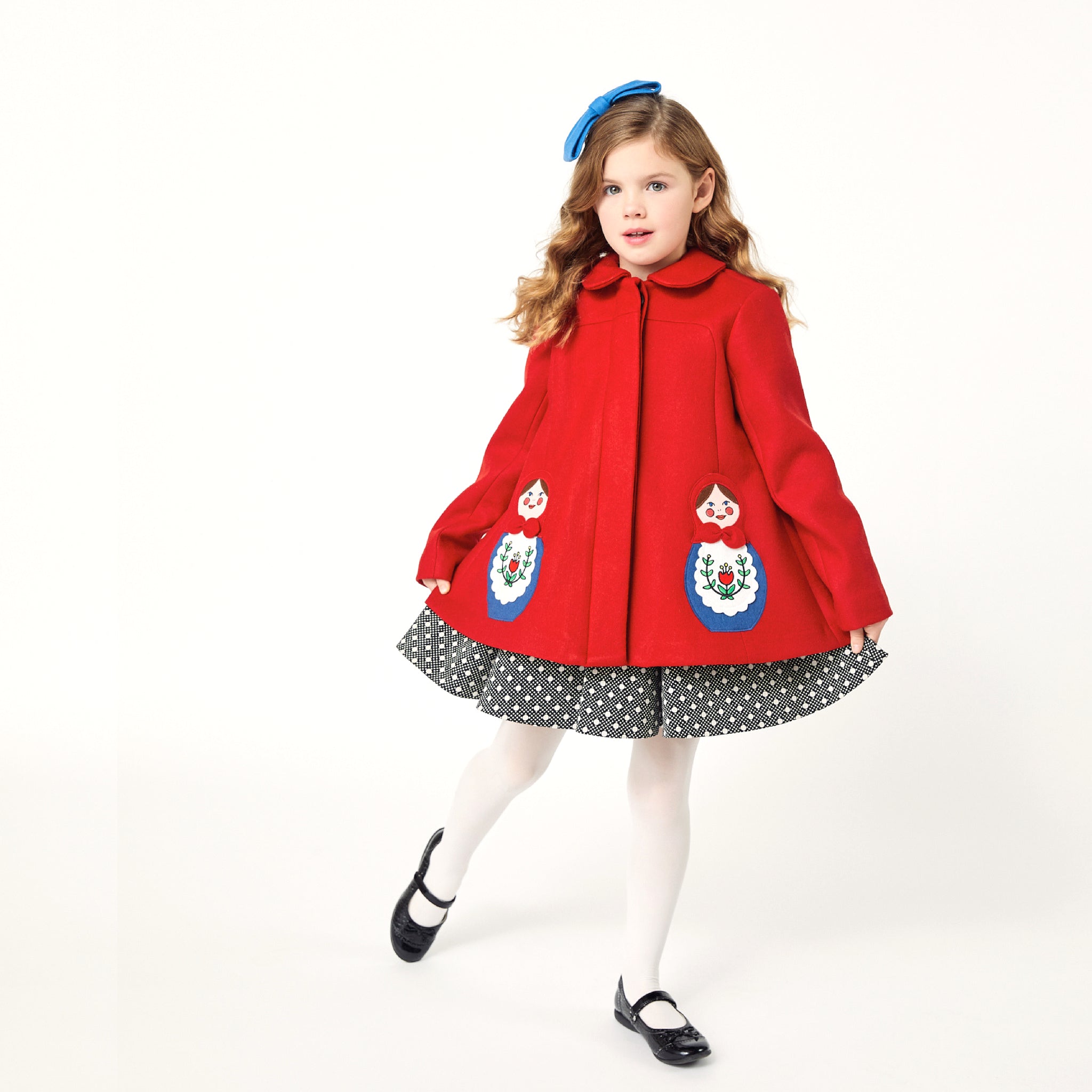 Girl modeling red coat while wearing a polkadot dress underneath and a blue bow on top of her head.