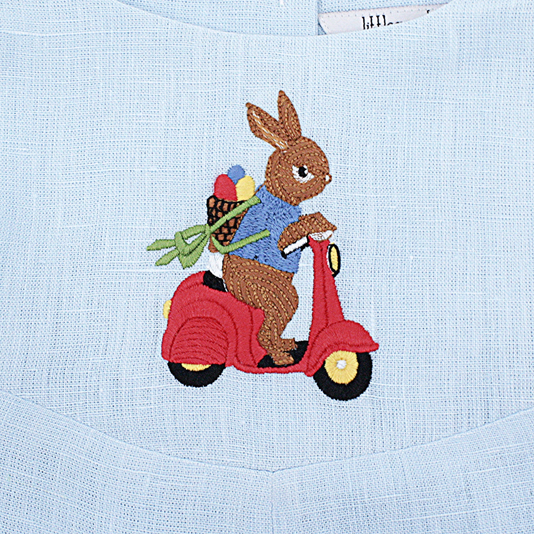 Close-up of embroidered bunny riding a red scooter on light blue linen romper. Detailed heirloom-quality stitching.