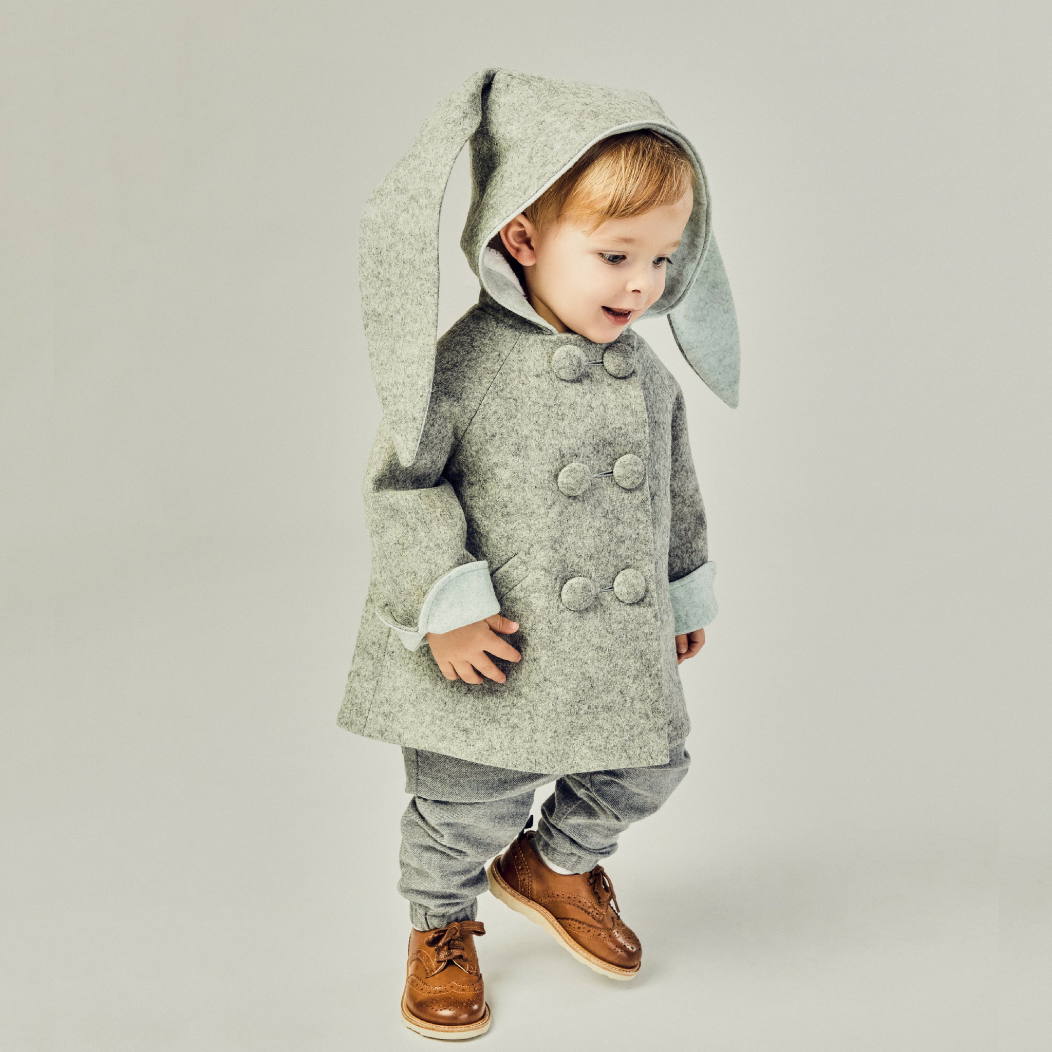 Boy wearing grey bunny coat with cuffed sleeves and hood up. One ear shows the grey exterior, the other reveals the light blue lining. Cozy and stylish