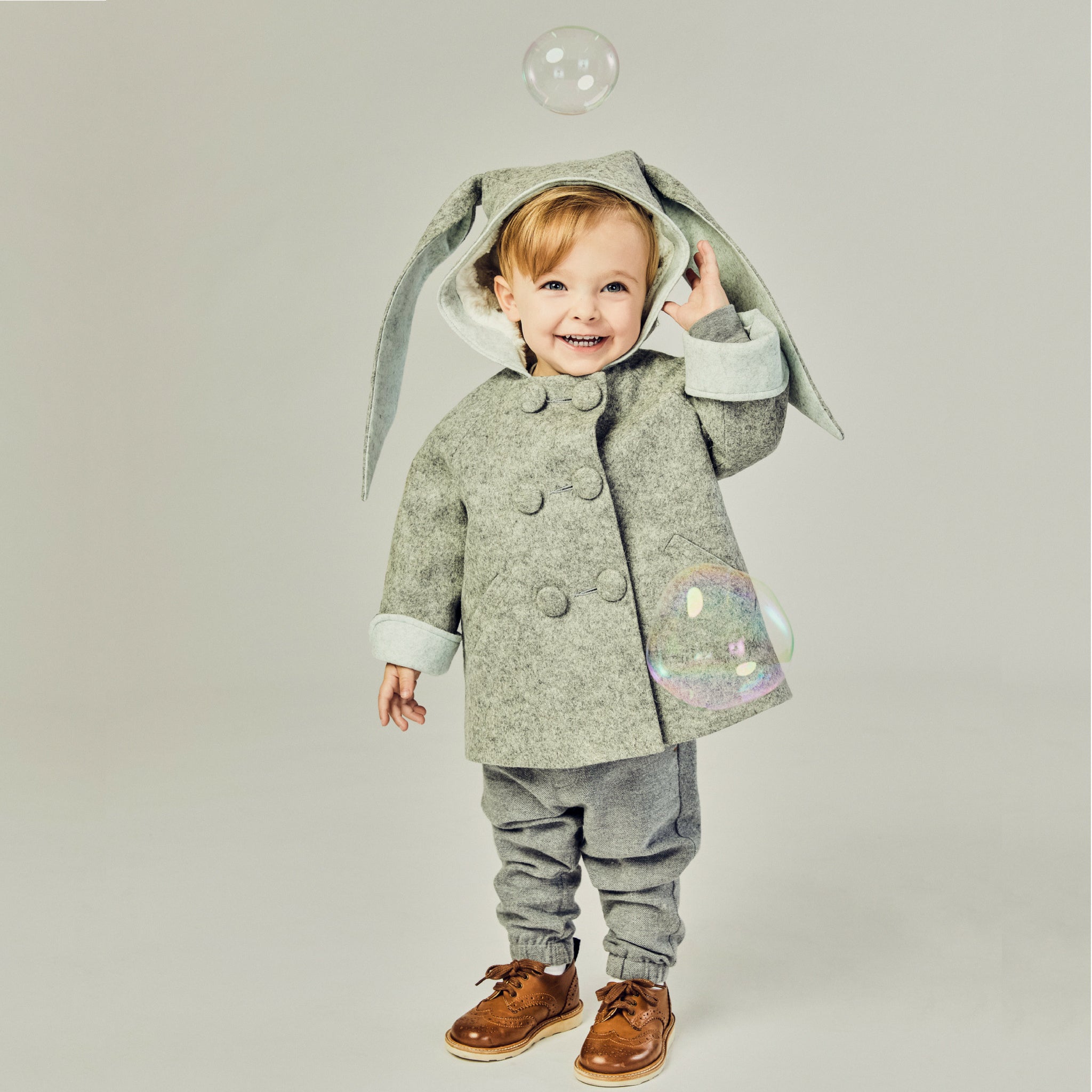 Smiling boy wearing grey bunny coat with hood up, revealing plush lining. One ear flops down while the other is lifted. Double-breasted buttons 