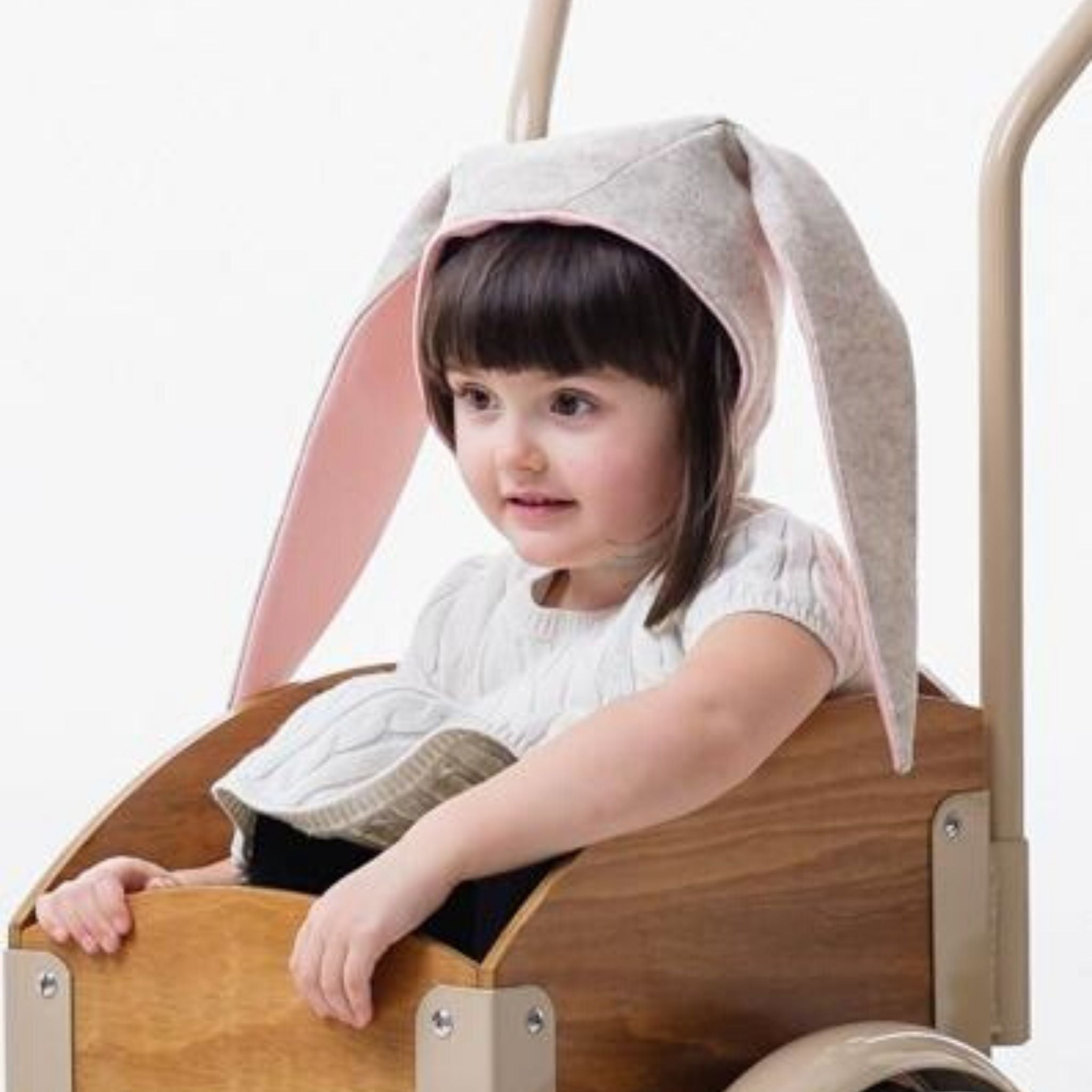 A child dressed in white, wearing Little Goodall's grey Snowshoe Rabbit Bunny Hat with soft floppy ears, sitting in a wooden wagon