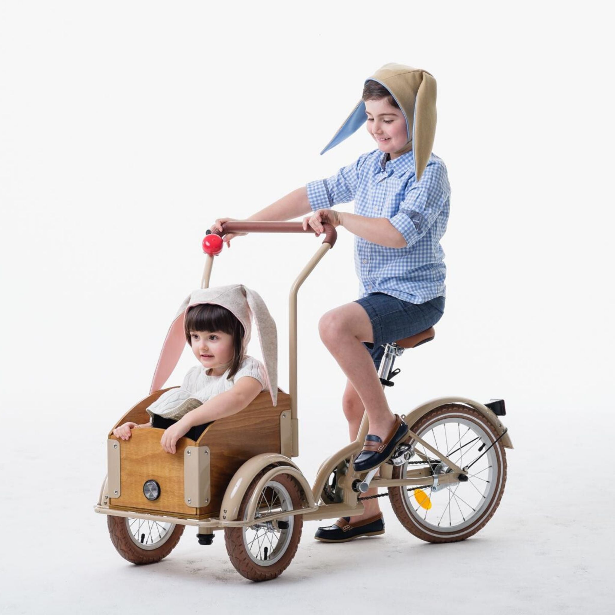 Two children on a tricycle, with one wearing the Snowshoe Rabbit Bunny Hat, grey with floppy ears