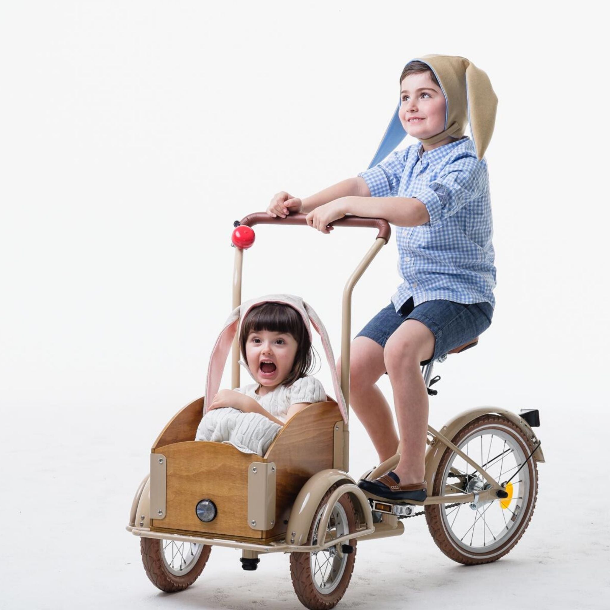 Two children on a tricycle, with one wearing a grey bunny hat featuring floppy ears