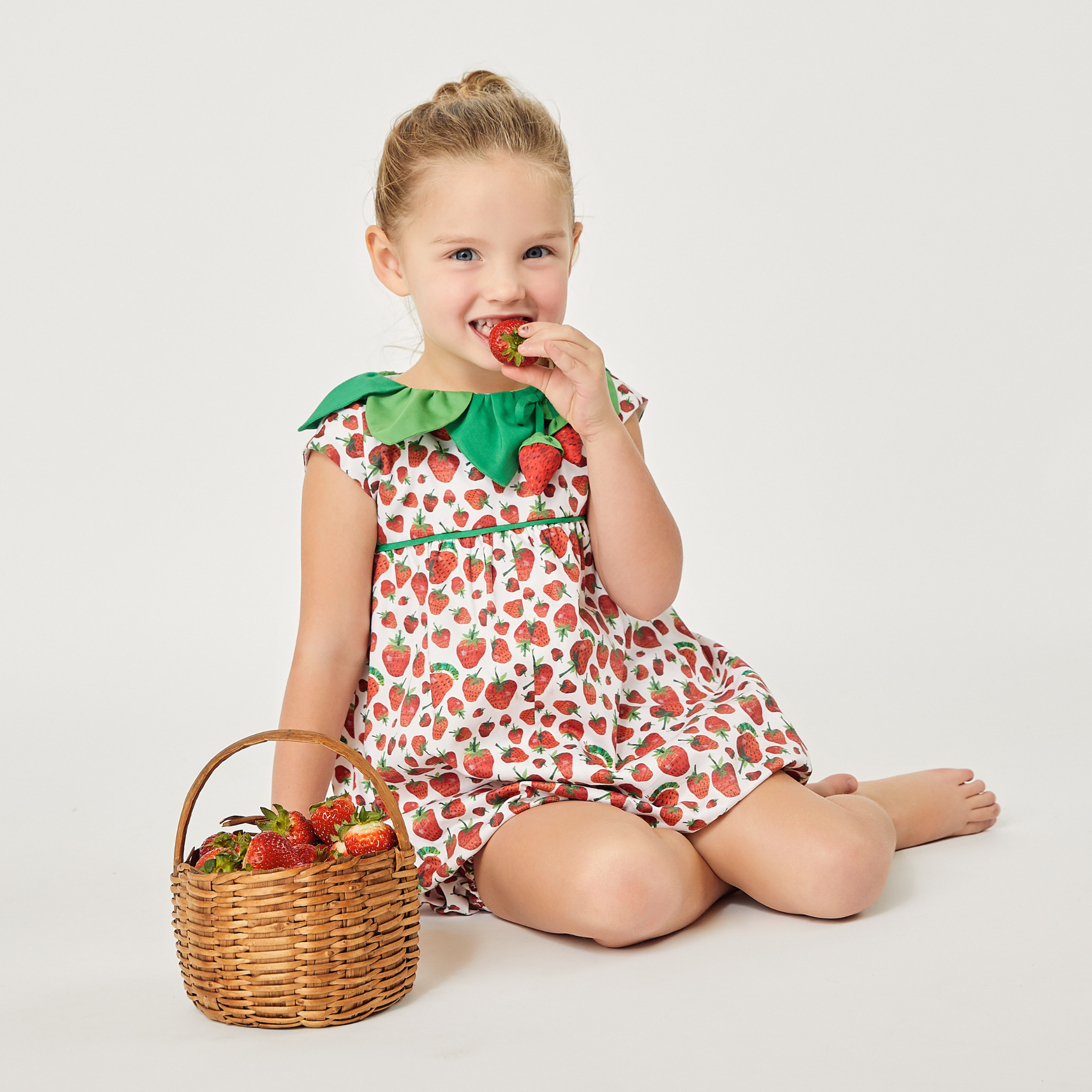 Toddler girl wearing a strawberry-print romper with a green leaf collar, sitting with a basket of strawberries 