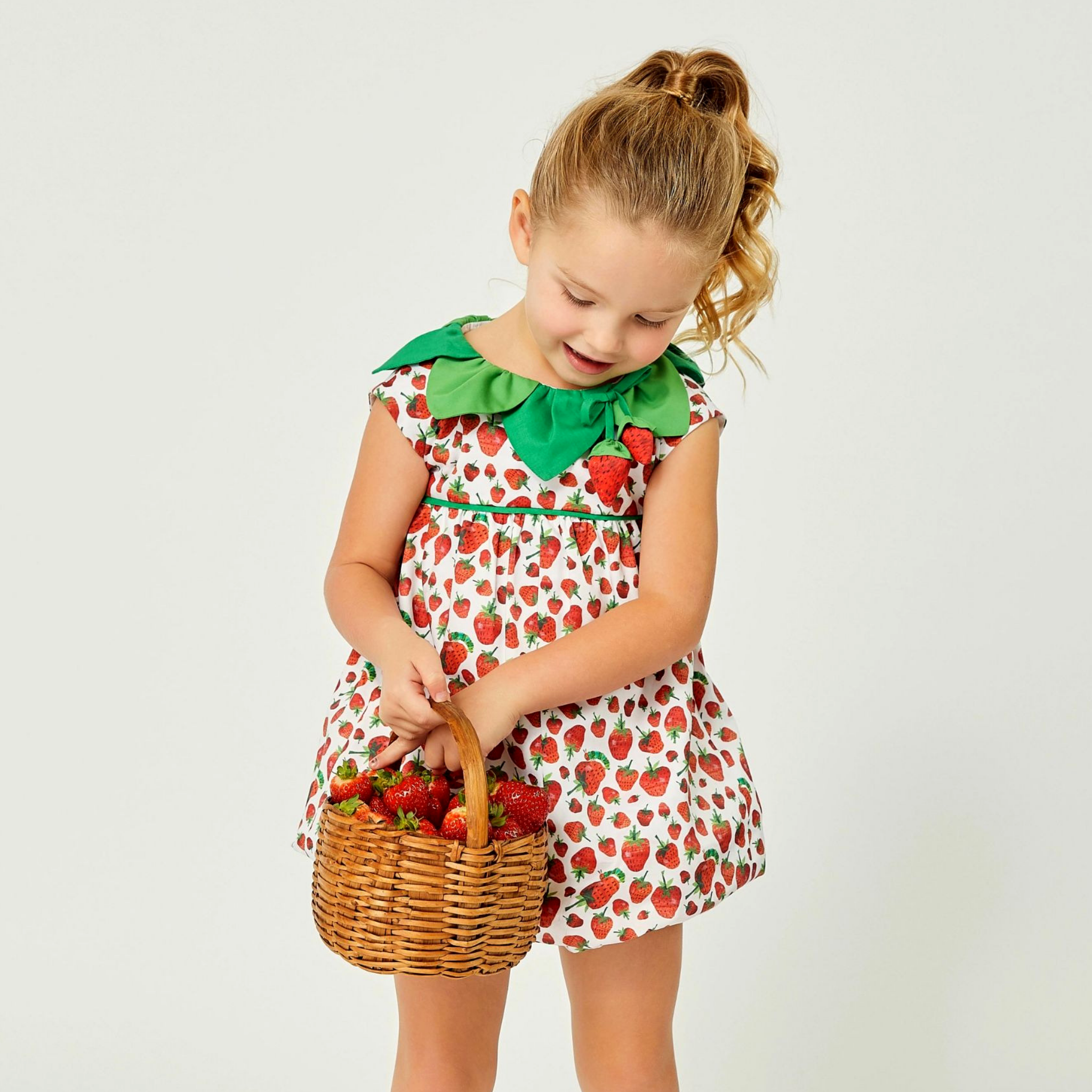 Toddler girl in a strawberry-print romper with a green leaf collar, holding a wicker basket filled with fresh strawberries.