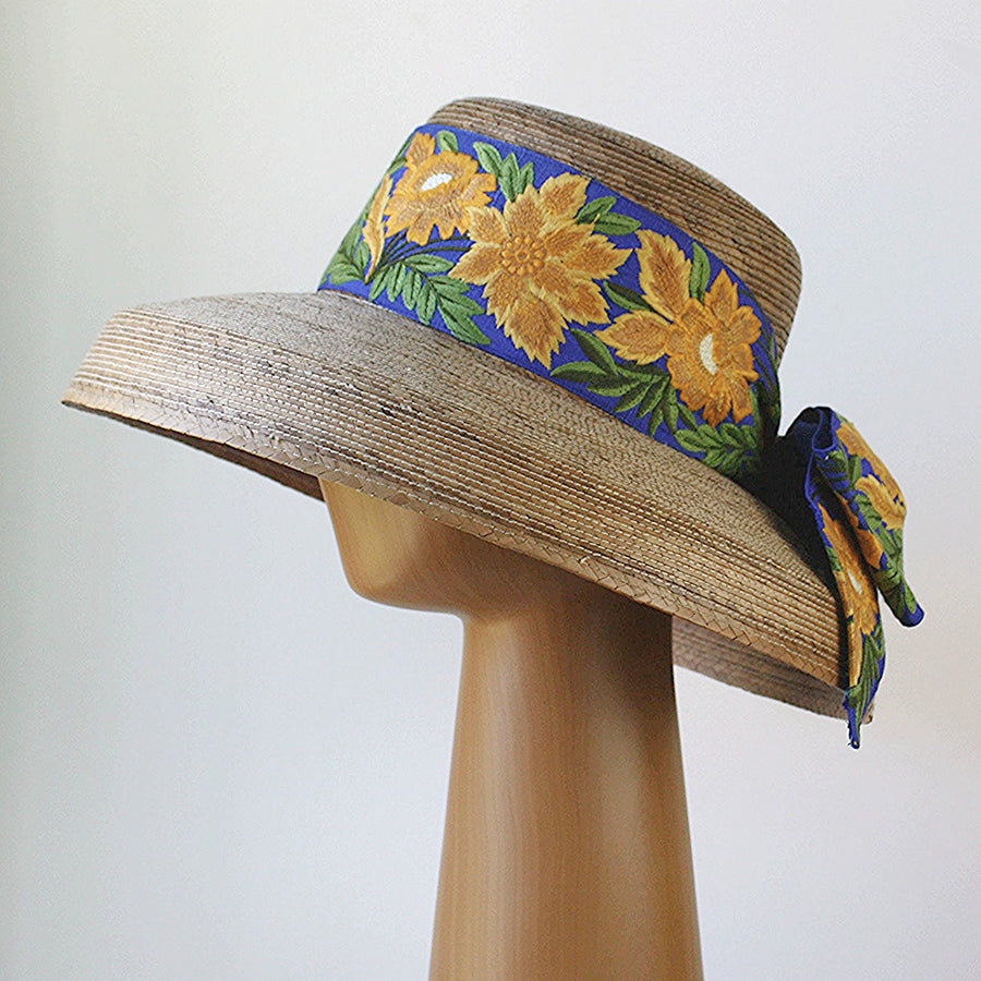 Profile view of Audrey Sunflower straw hat with wide brim and French blue ribbon featuring yellow floral embroidery and tied bow