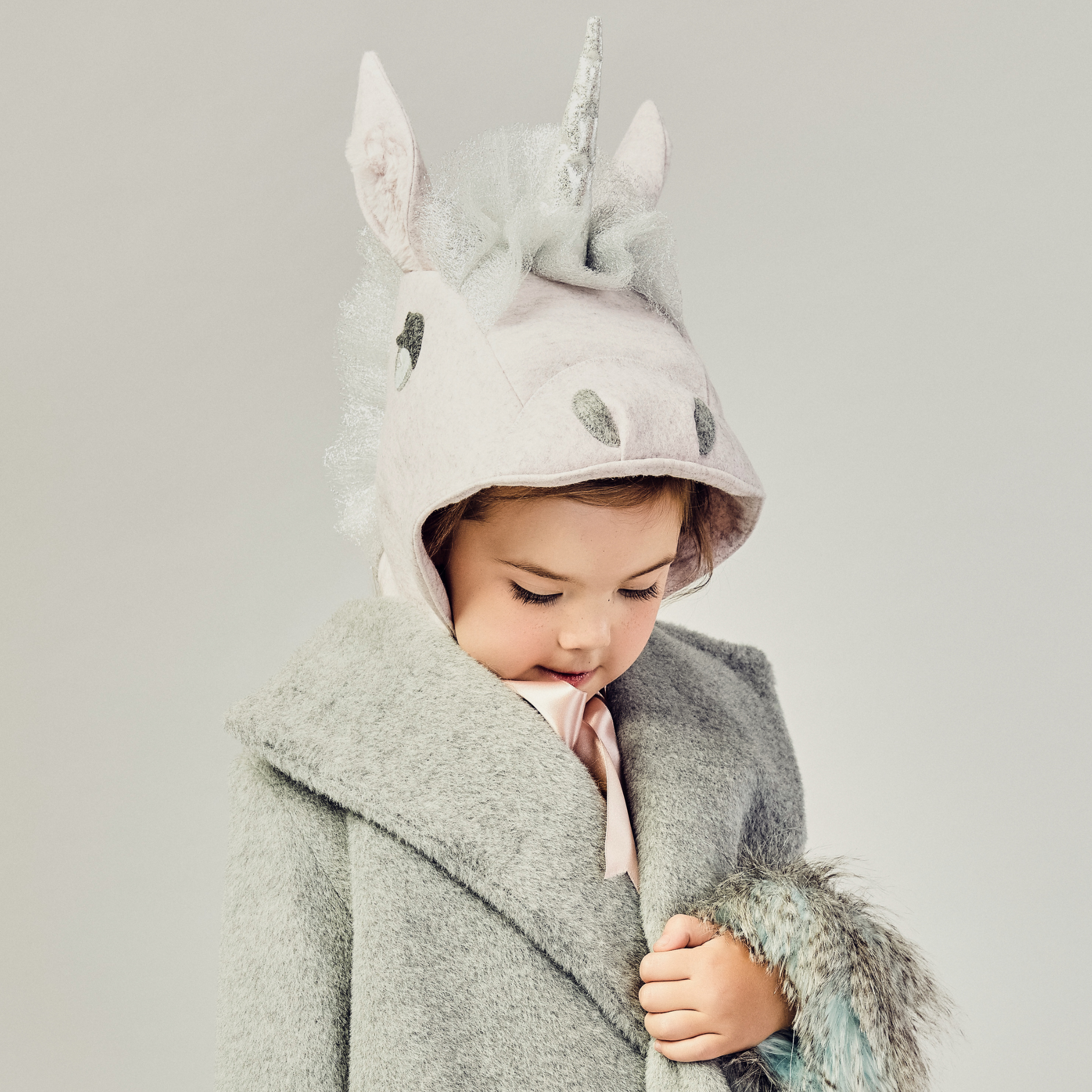 Young child wearing a pale pink wool unicorn hat with a silver mane and satin ribbon ties, bundled up in a cozy grey coat with faux fur trim