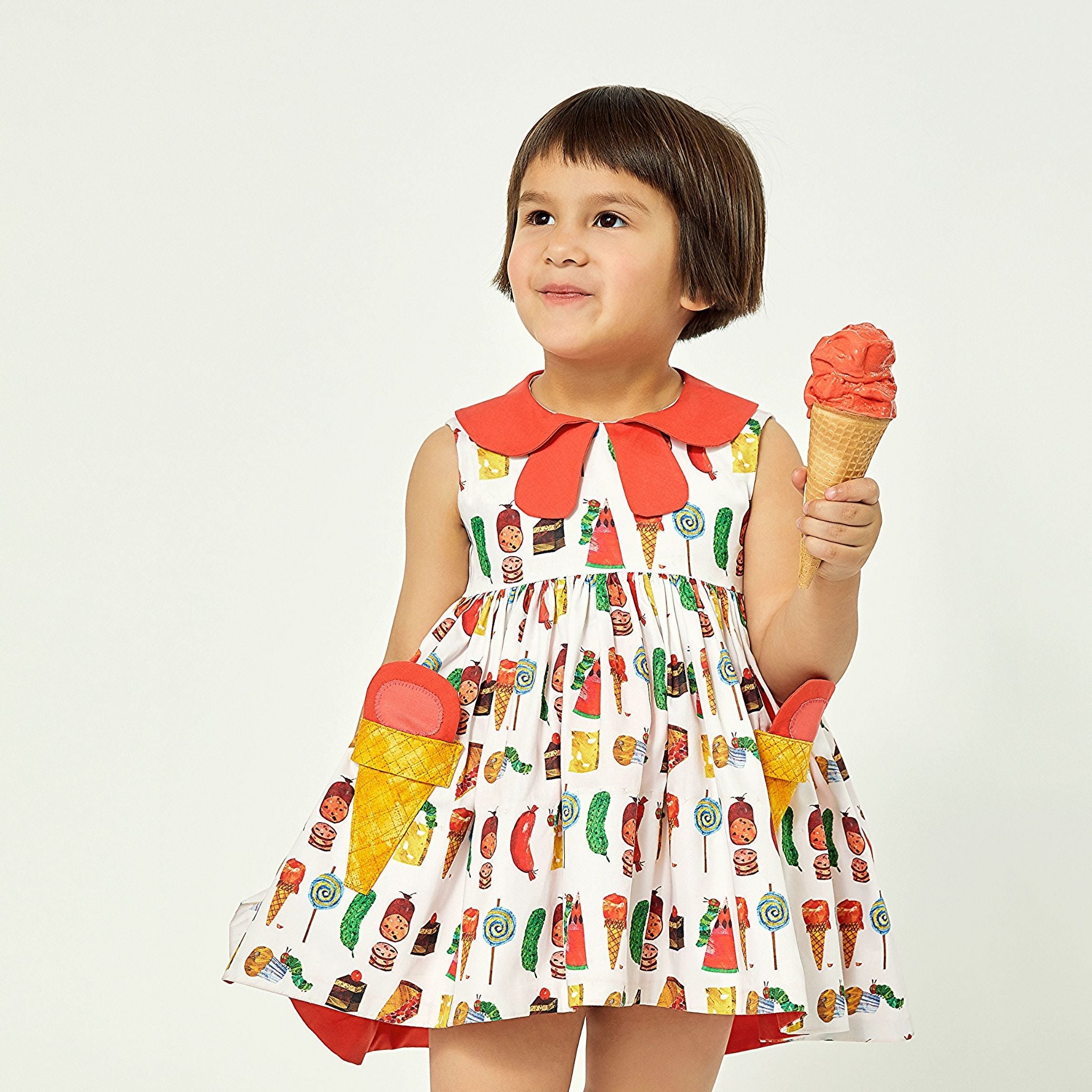 Girl wearing cotton Two Scoops Dress with ice cream pockets and a coral collar, holding an ice cream cone, featuring a fun candy-themed print