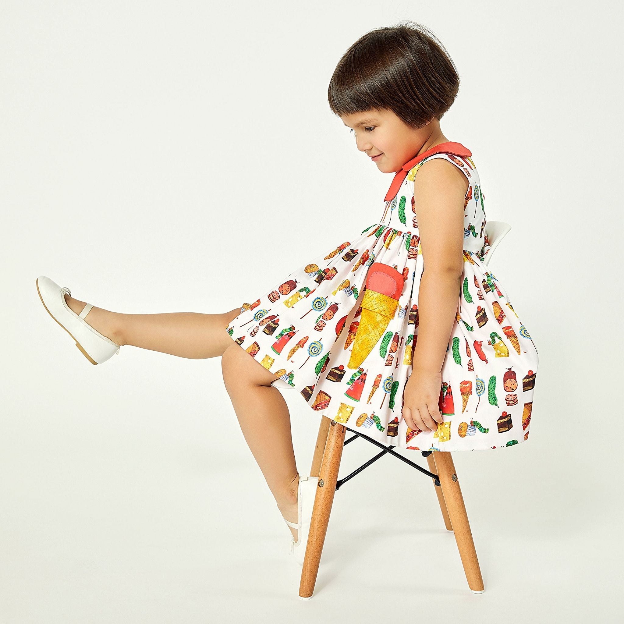 Girl in cotton Two Scoops Dress with The Very Hungry Caterpillar print, coral collar, and quilted ice cream pockets, sitting on chair with playful pose