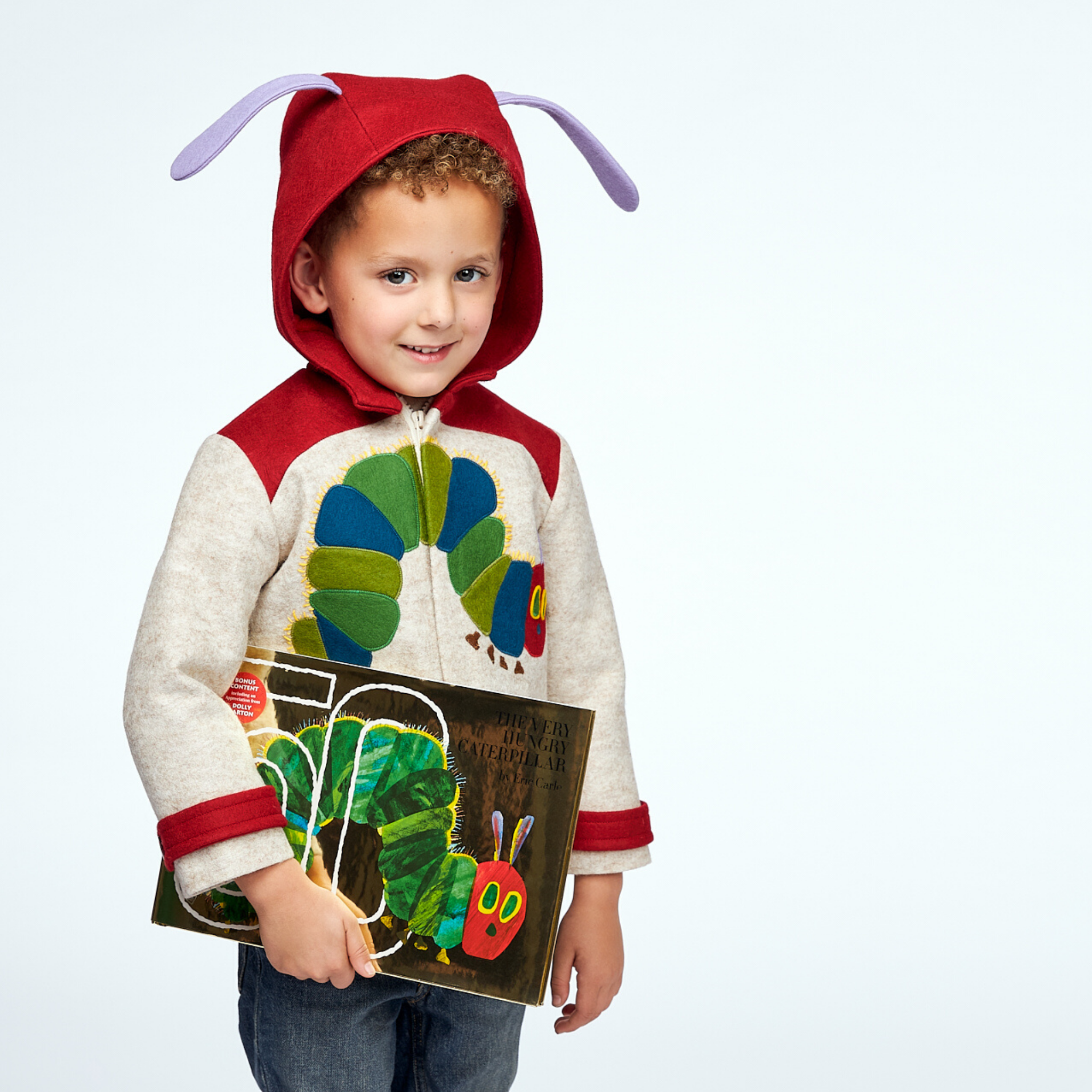 Smiling child wearing a Hungry Caterpillar wool coat with a red hood and antennae, holding the matching book, perfect for toddlers and kids.