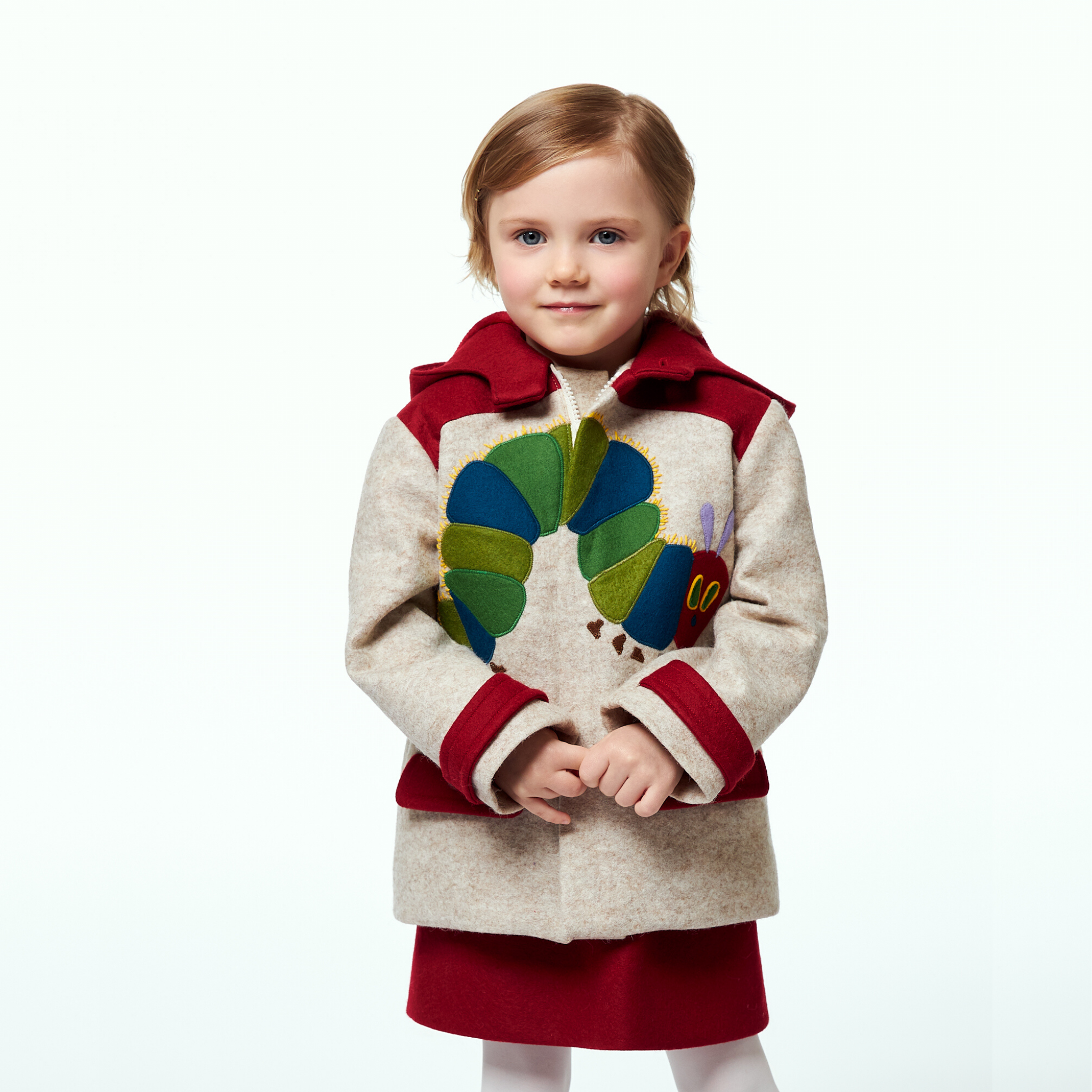 A young girl wearing a Hungry Caterpillar wool coat featuring a whimsical caterpillar applique, red collar, and cuffs, paired with a red dress