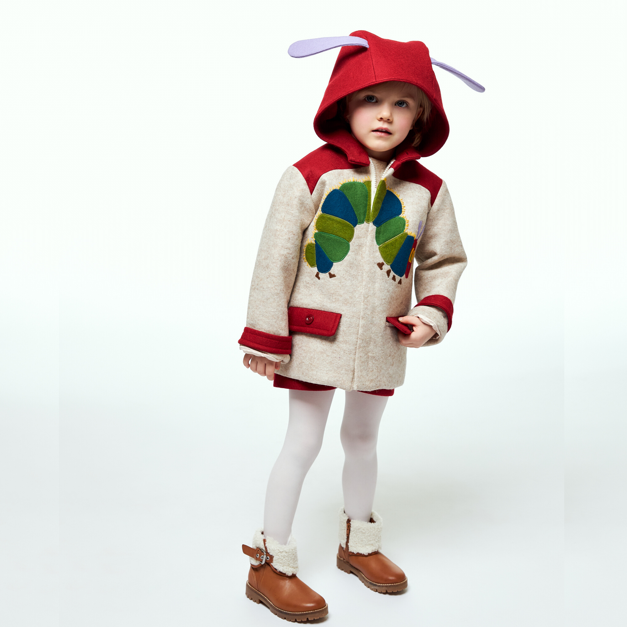 young girl wearing a Hungry Caterpillar wool coat with the hood up, featuring playful caterpillar details, red accents, and matching tights and boots