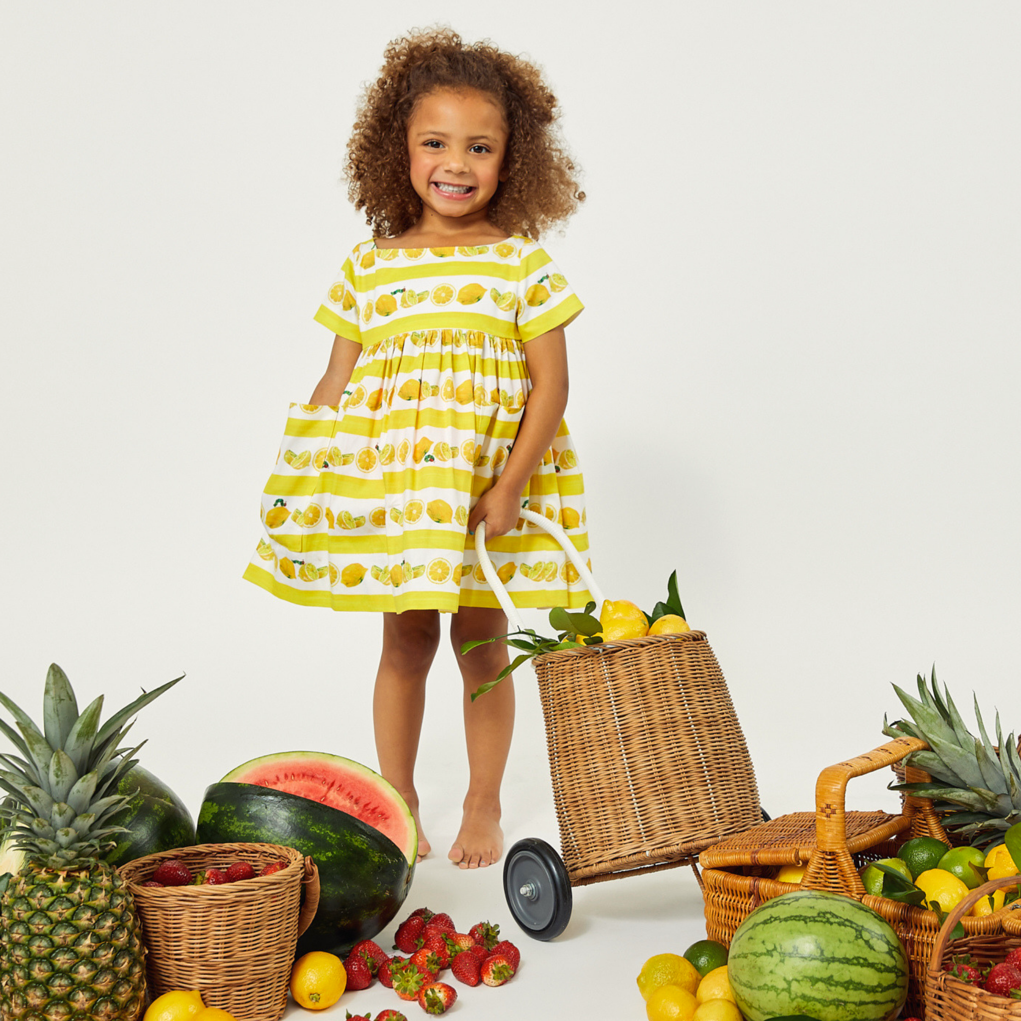 A young girl in a striped yellow dress with lemon patterns stands barefoot at a fruit market, holding a mini cart of lemons