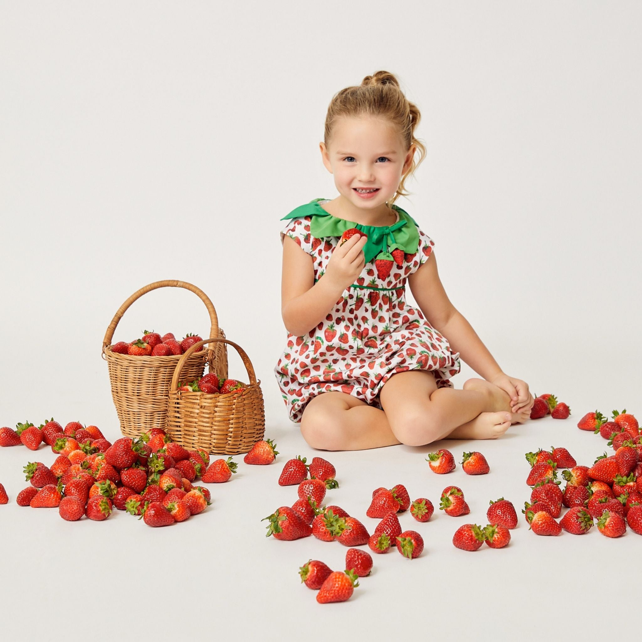 Smiling toddler wearing a strawberry-print romper, sitting with fresh strawberries and a wicker basket on a white background