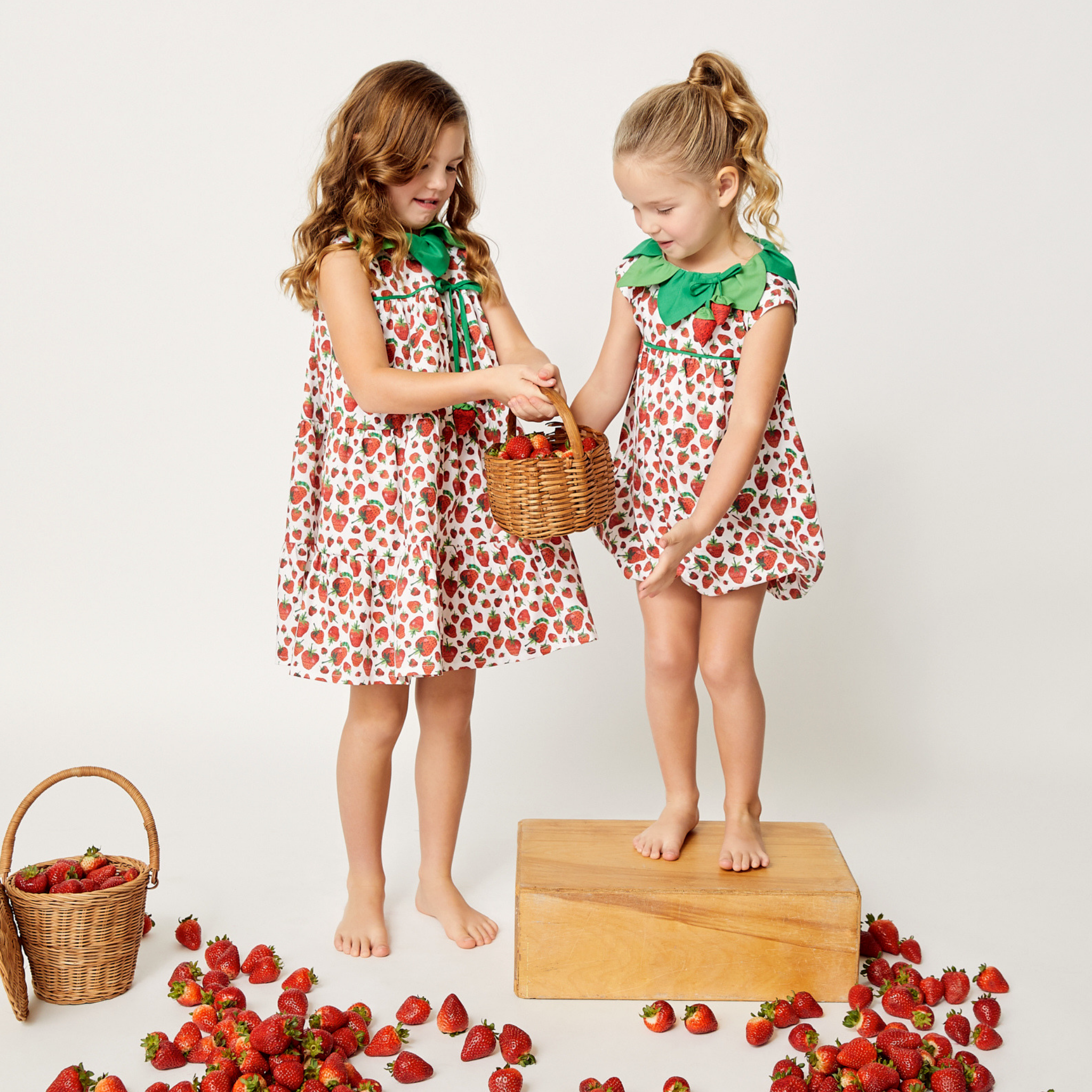 Toddler and girl in coordinated strawberry dresses and rompers, sharing a basket of strawberries