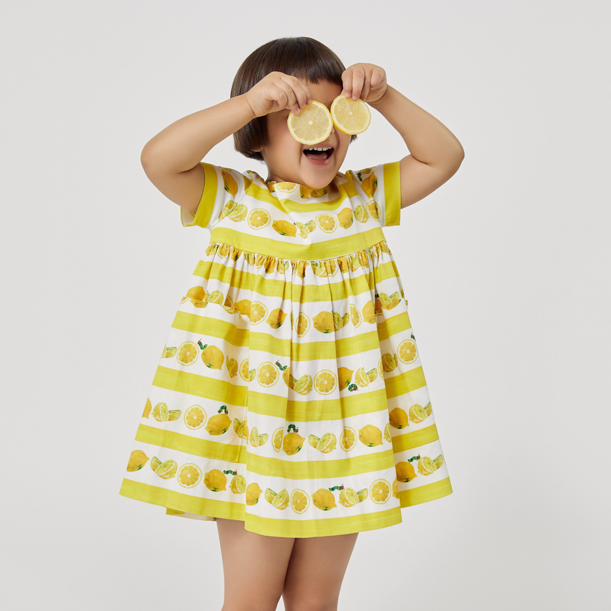 Smiling child in a lemon print dress holding lemon slices over her eyes, wearing The Very Hungry Caterpillar Lemonade Dress