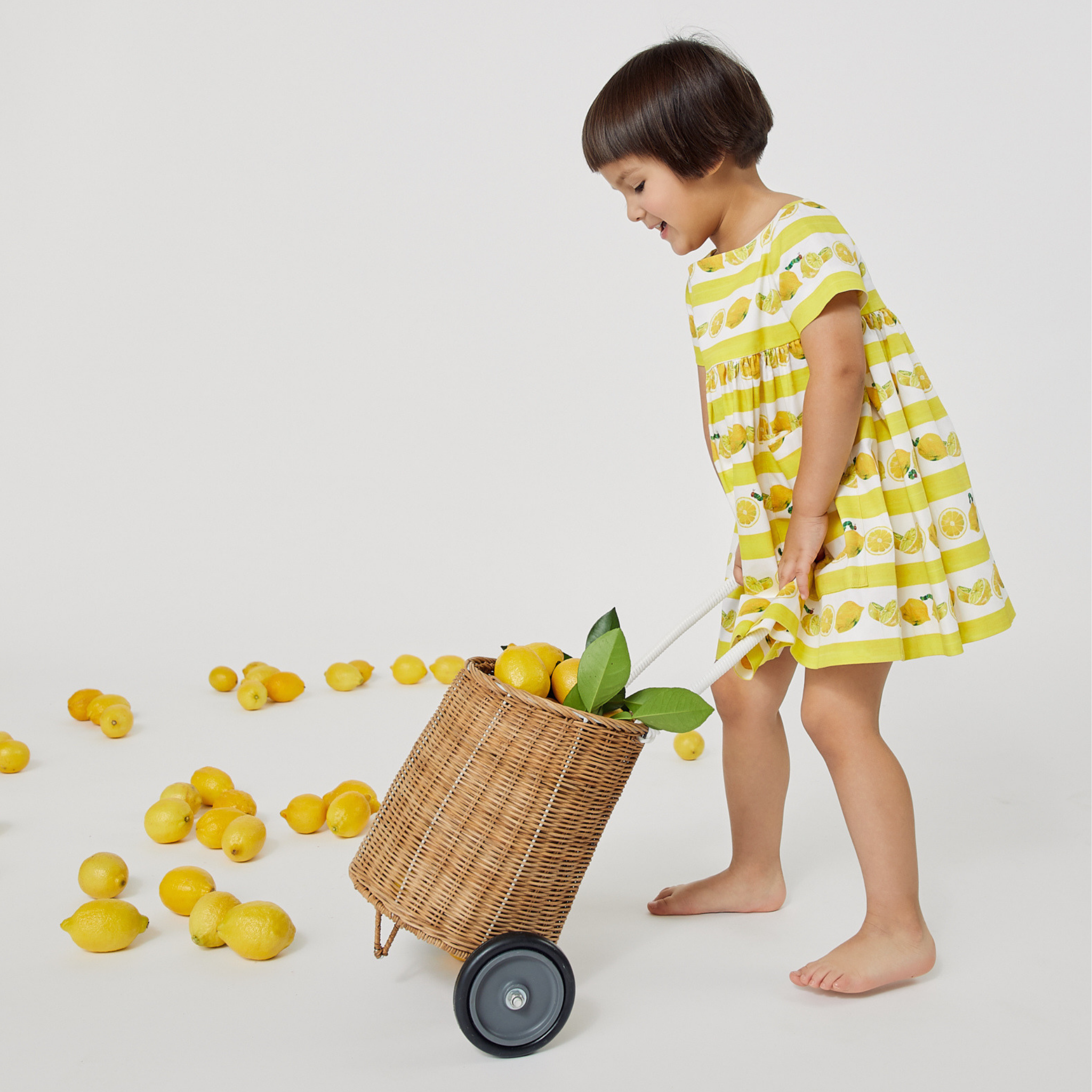 Young girl in a lemon-print dress playfully pulling a wicker cart filled with lemons, surrounded by scattered lemons.